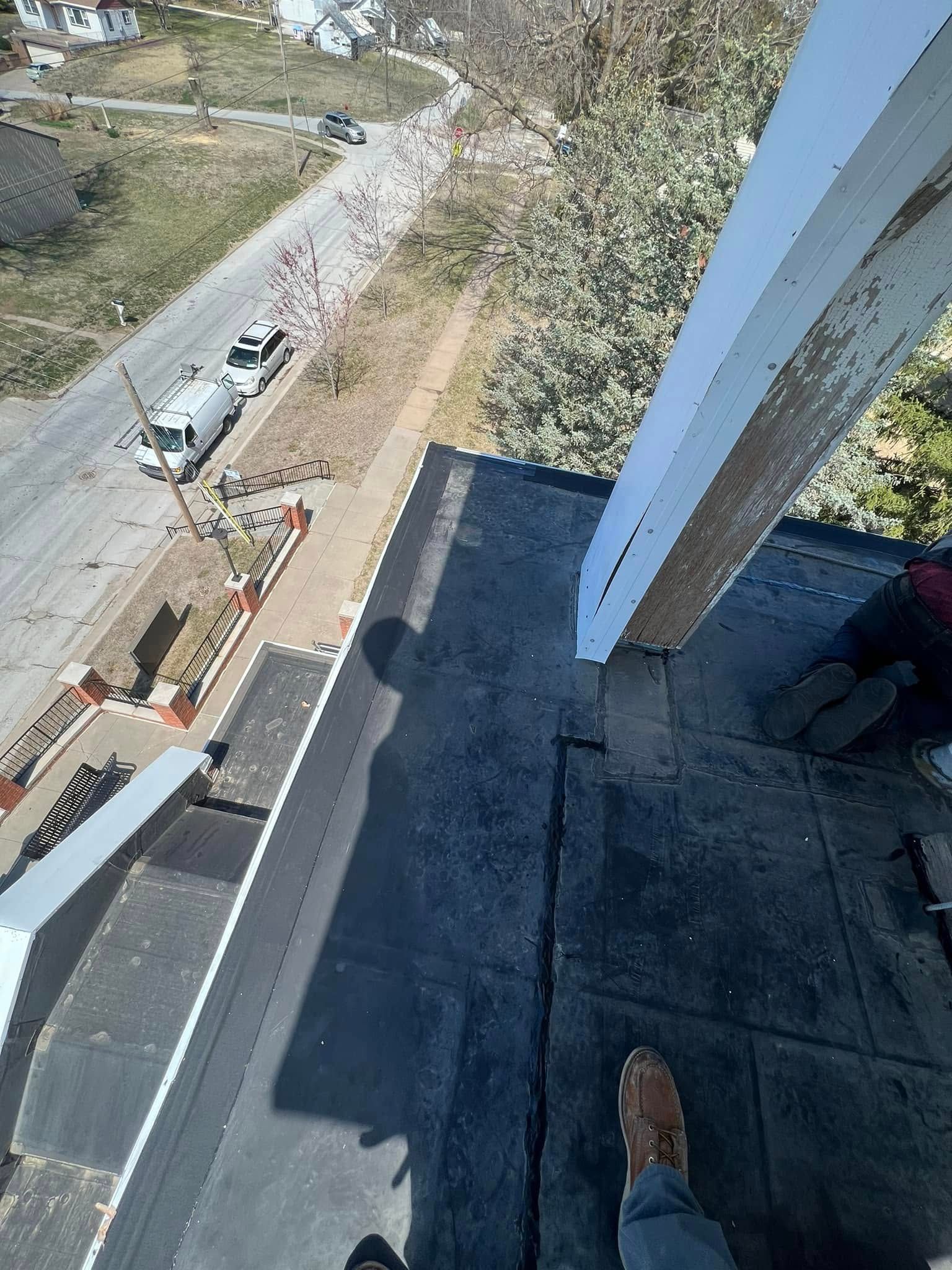 Rooftop edge view of a road, parked cars, and trees below, with a person’s shoe visible in the foreground.
