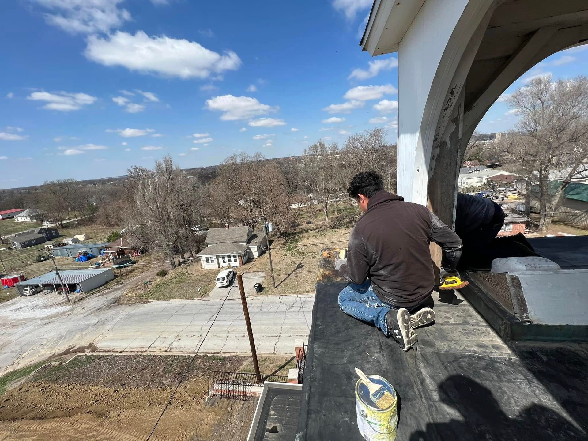 Two workers sit on a rooftop edge overlooking a wooded neighborhood on a clear day.