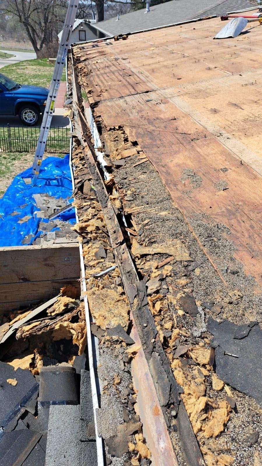 Severely damaged roof with exposed wood and torn shingles beside a blue tarp and house gutter.