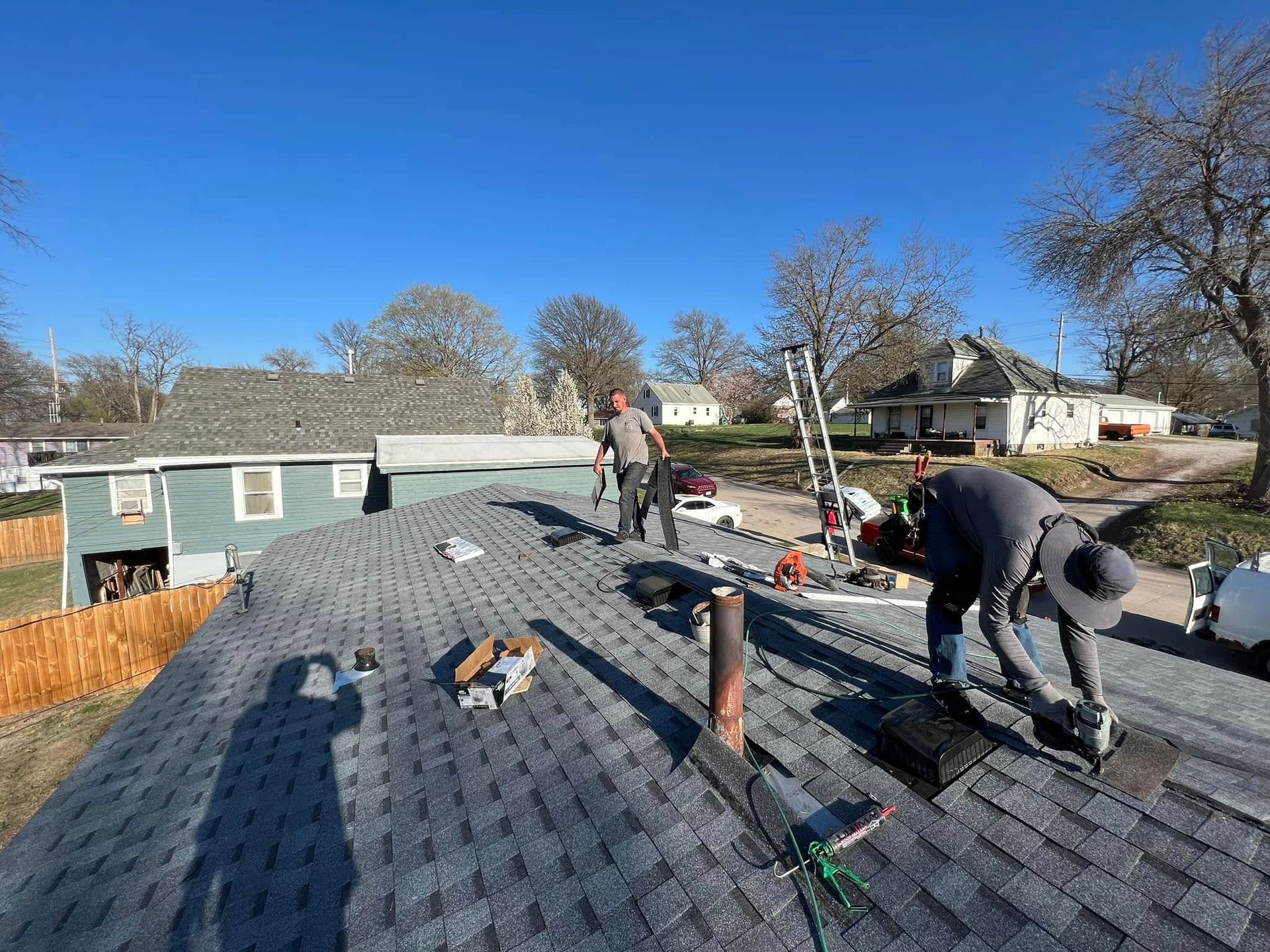 Workers installing shingles on a roof under a clear blue sky, with houses and trees in the background