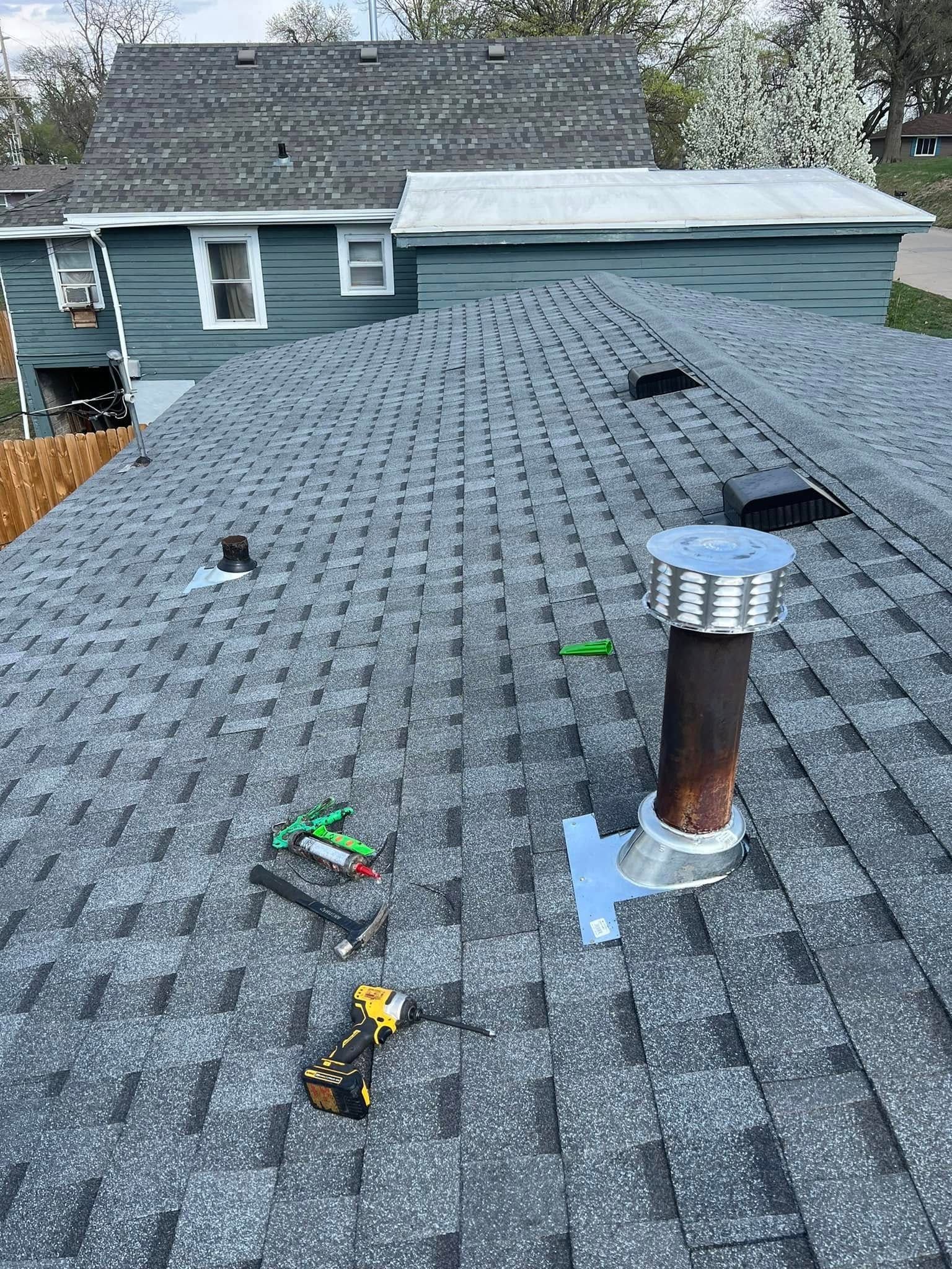 Roof shingles with a vent pipe and small tools on a sloped gray rooftop, house in background