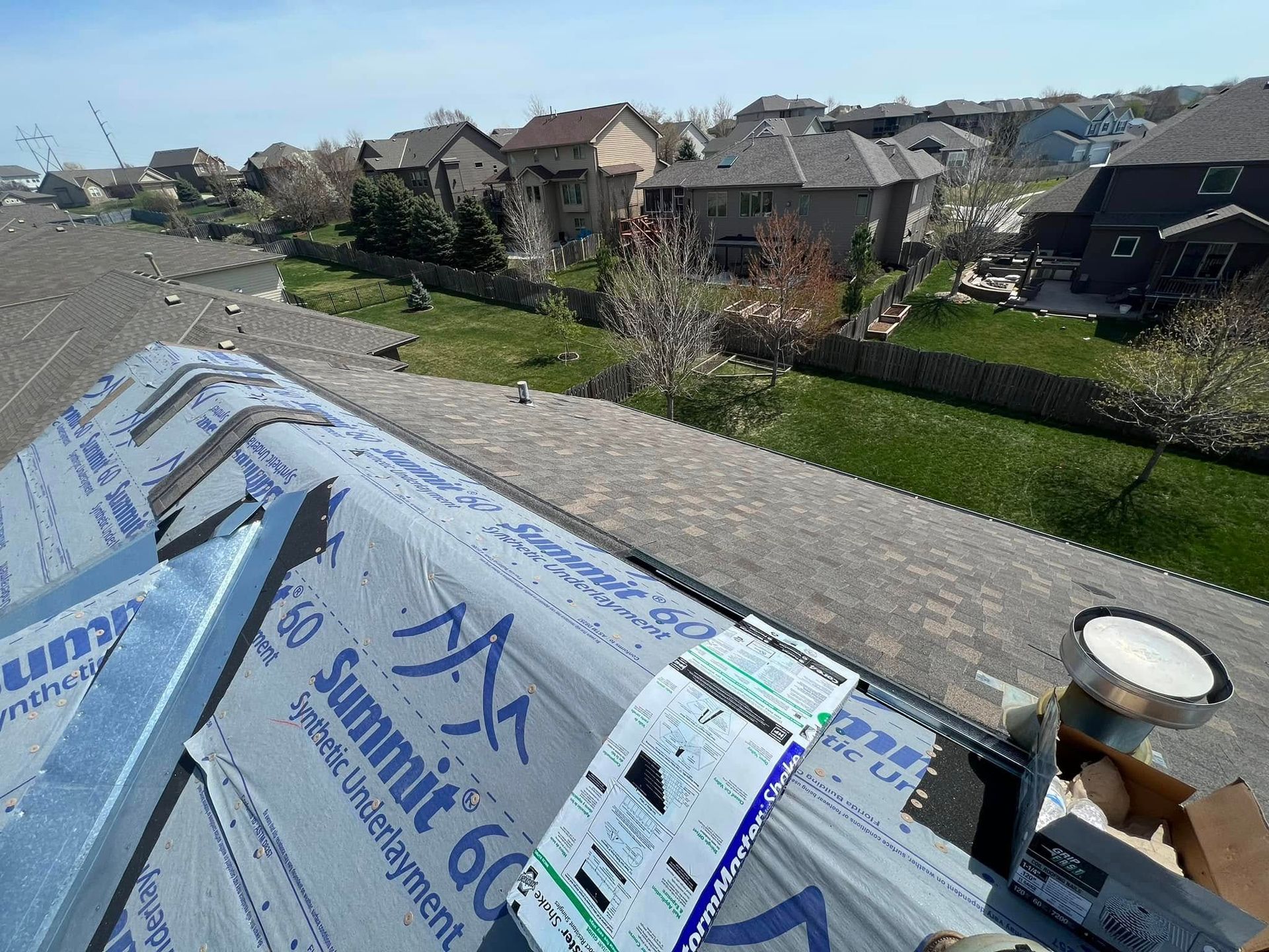 Roofer’s view of blue underlayment and shingles on a residential roof with houses and lawn below