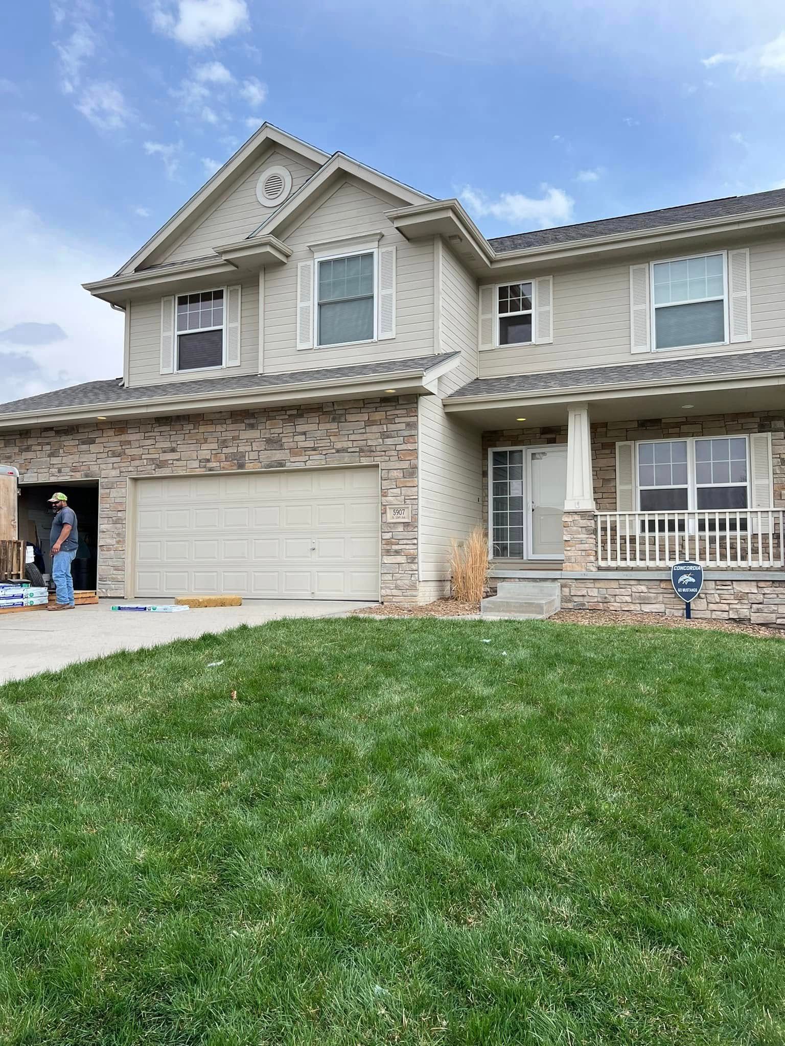 Two-story suburban stone house with white garage, front porch, and green lawn under a blue sky.