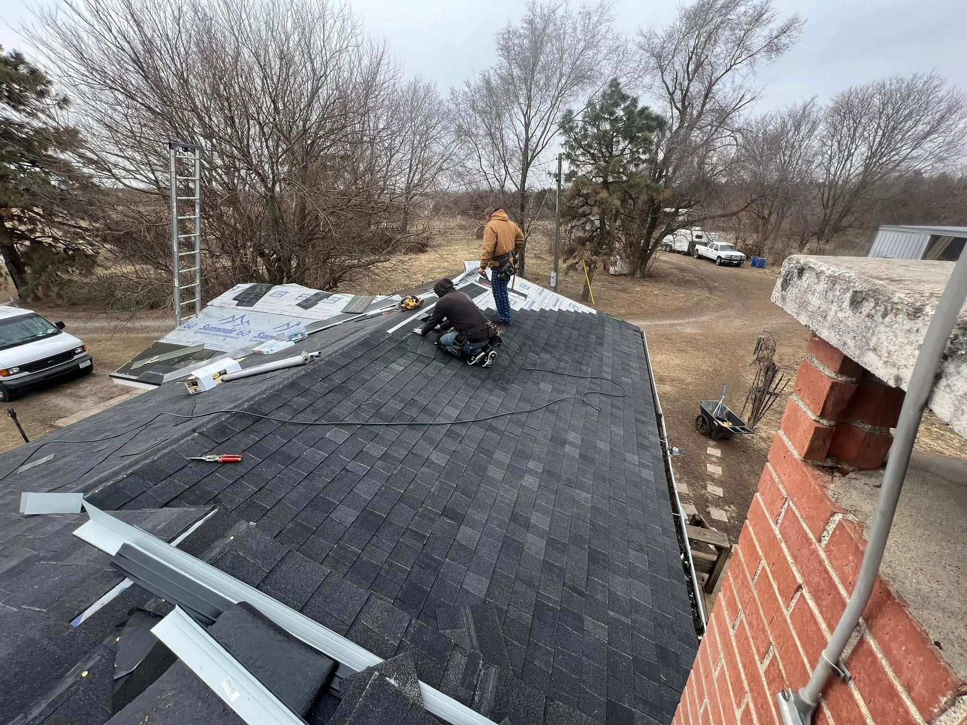 Worker on a roof inspecting shingles near a brick chimney on a cloudy day.