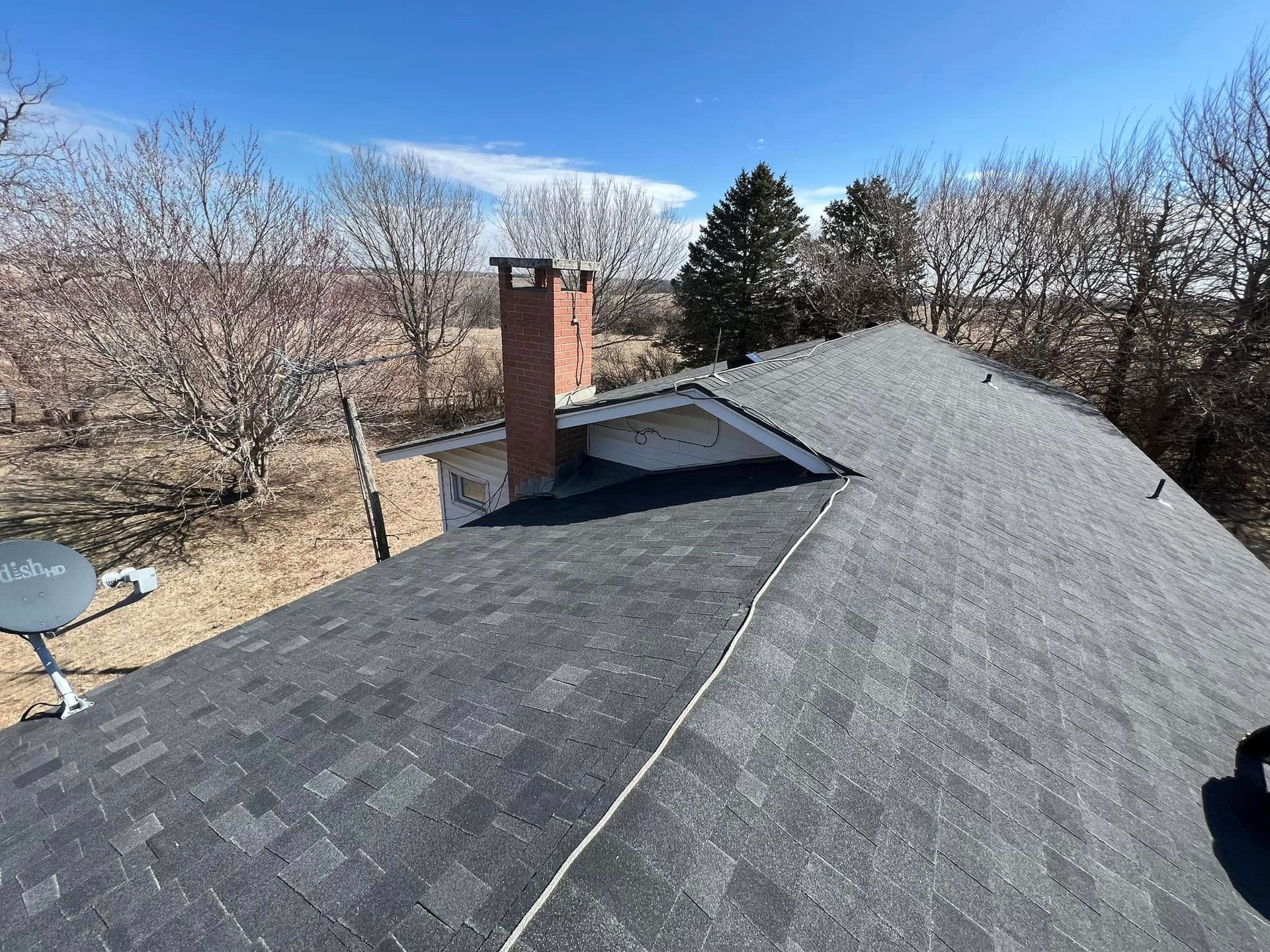 Rooftop with dark gray shingles, brick chimney, satellite dish, and leafless trees under a blue sky