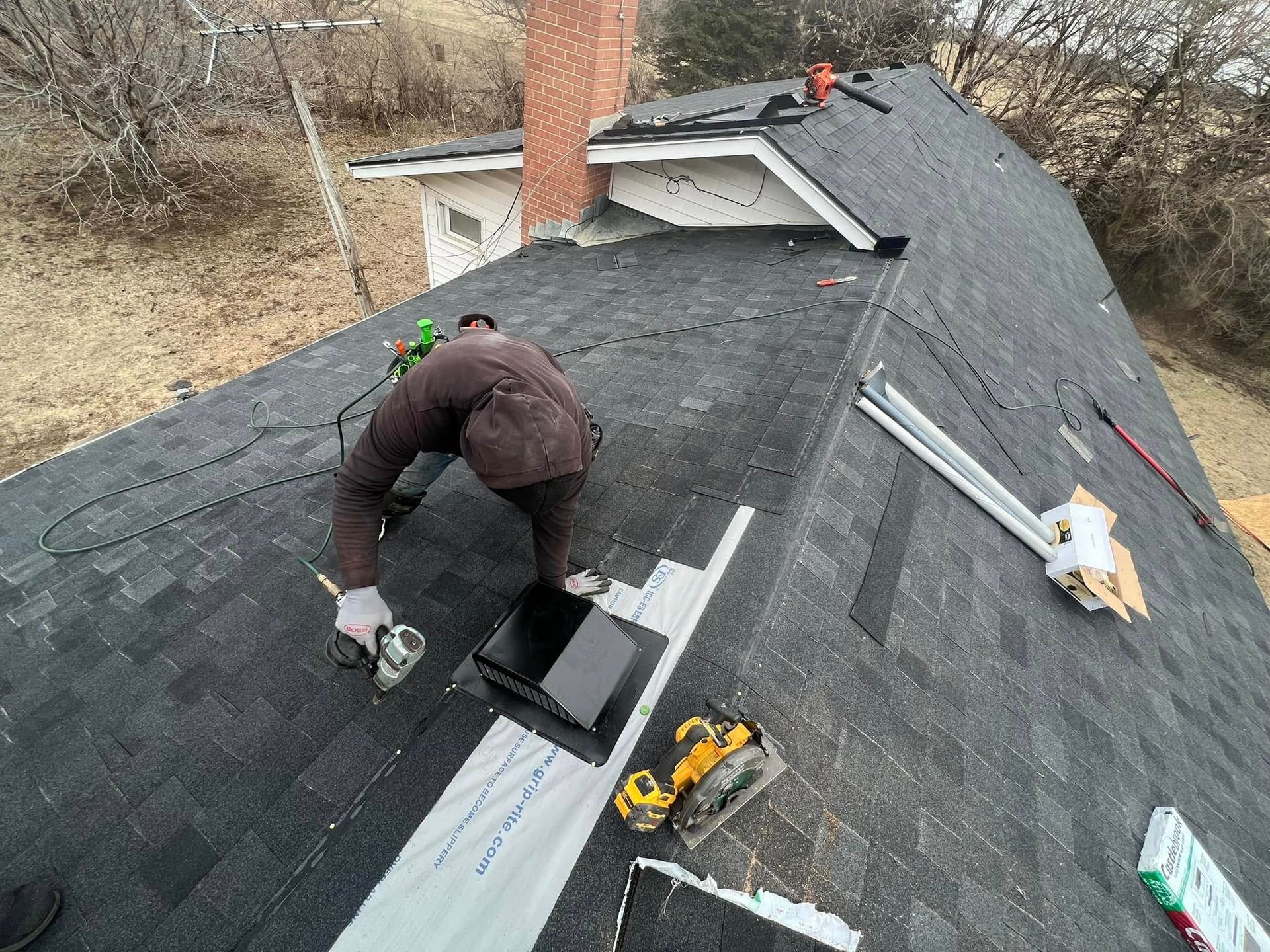 Worker repairing a black shingle roof with tools near a chimney.