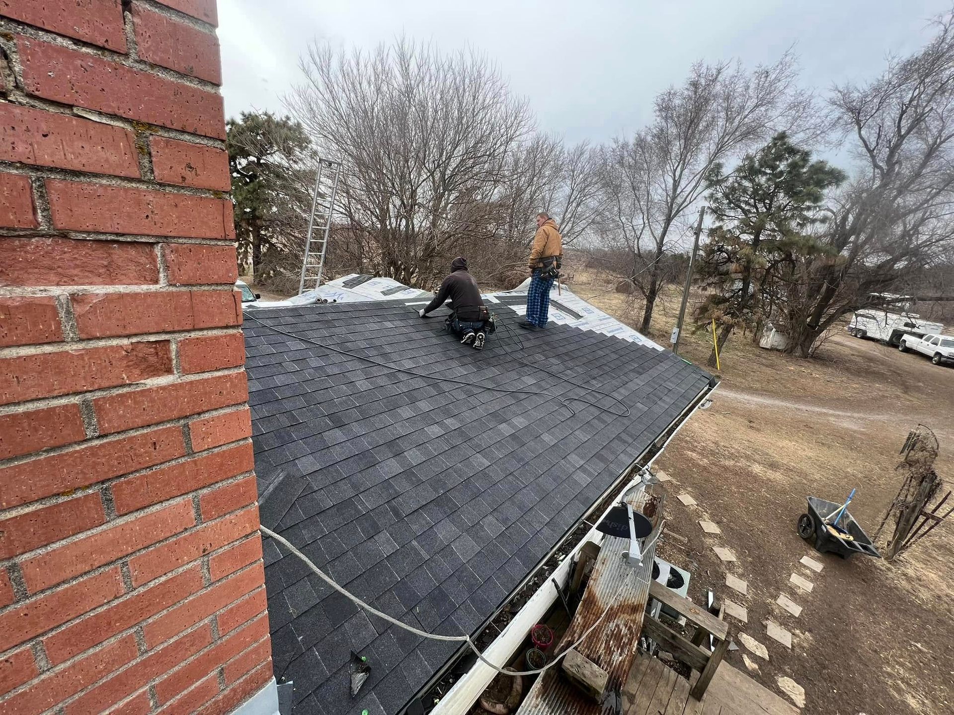 Workers installing shingles on a sloped roof beside a brick chimney and bare trees
