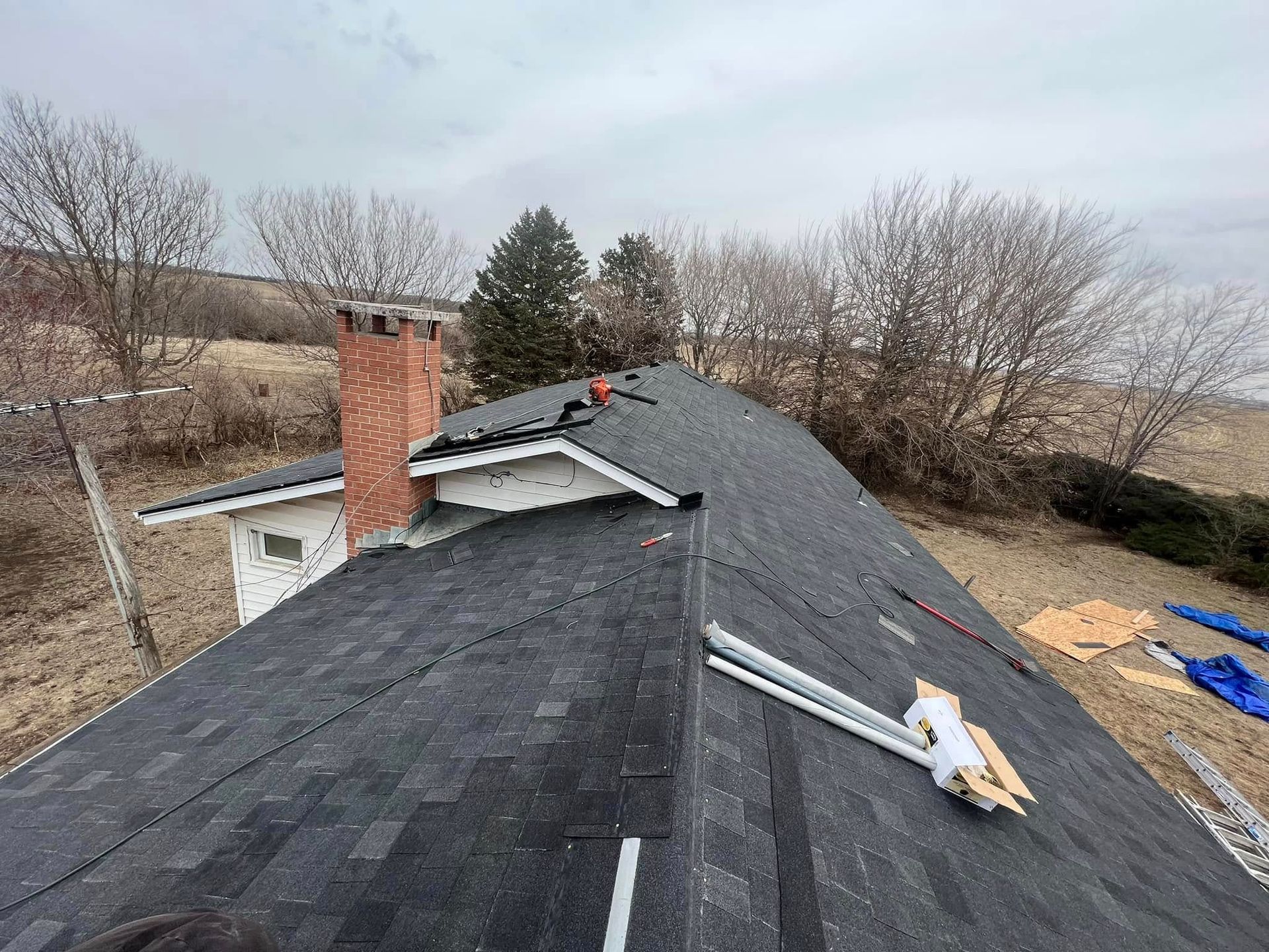 Roofer on a shingled roof beside a brick chimney, working near a leafless yard.
