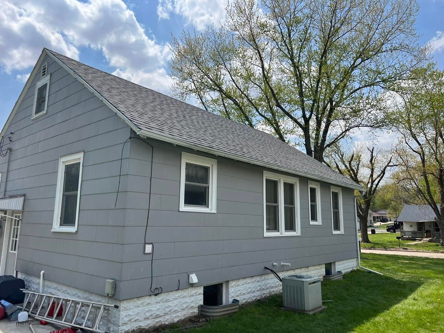 Gray house exterior with white trim, side yard, and two trash bins under a tree.