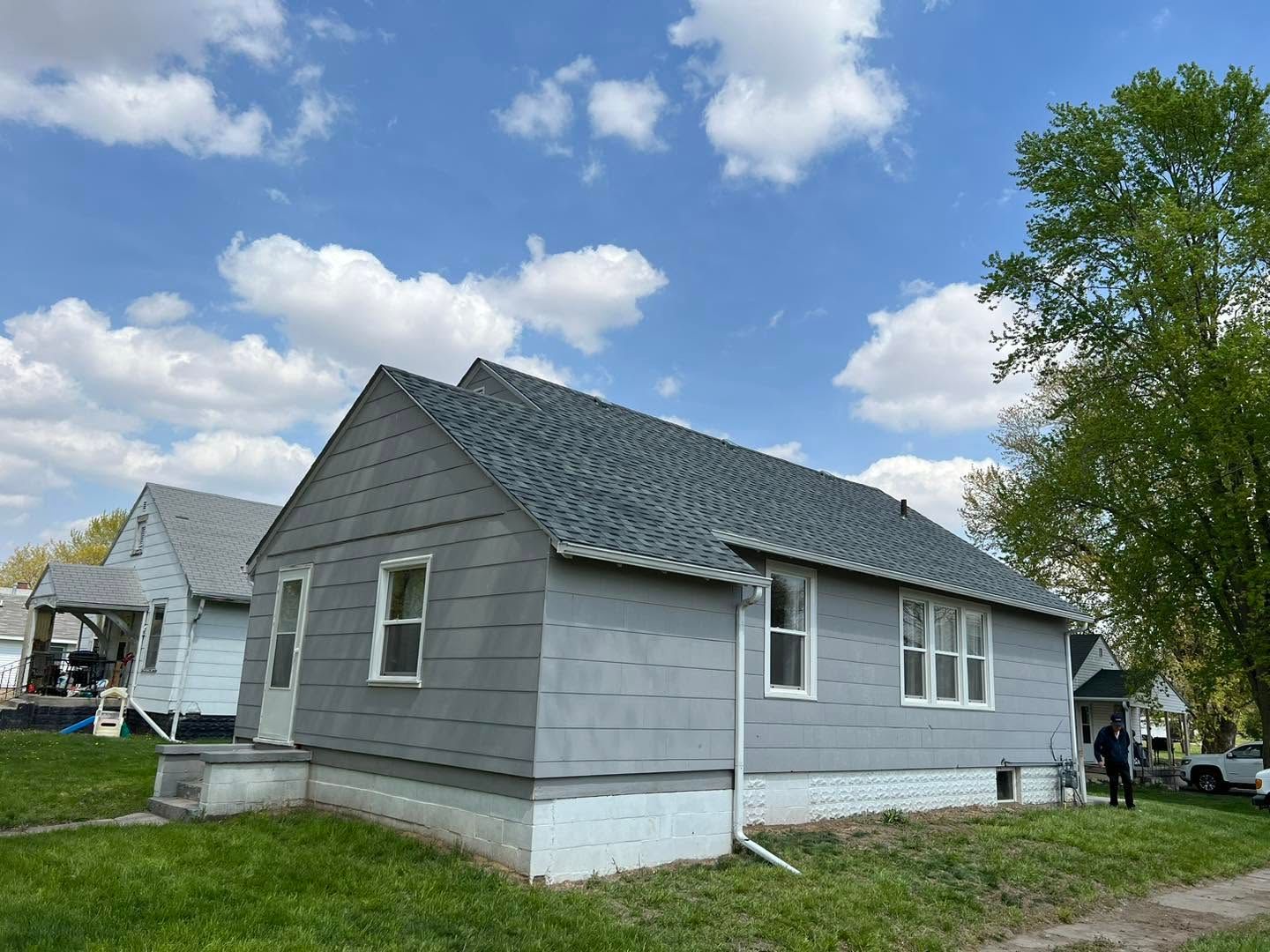 Gray house with white trim and sloped roof on a sunny day, with another house and trees nearby