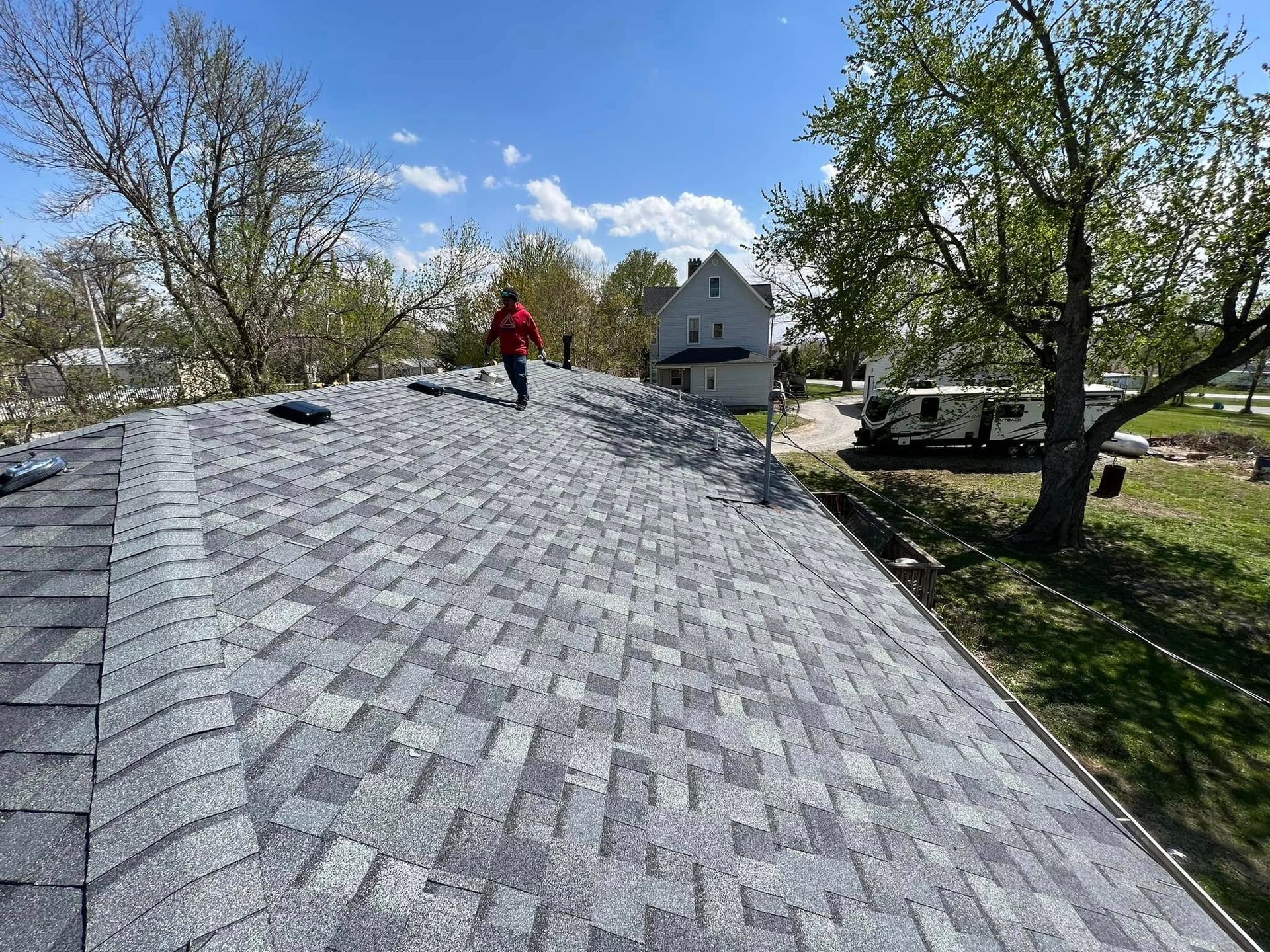 Worker on a shingled roof inspecting installation, with houses and trees in the background.