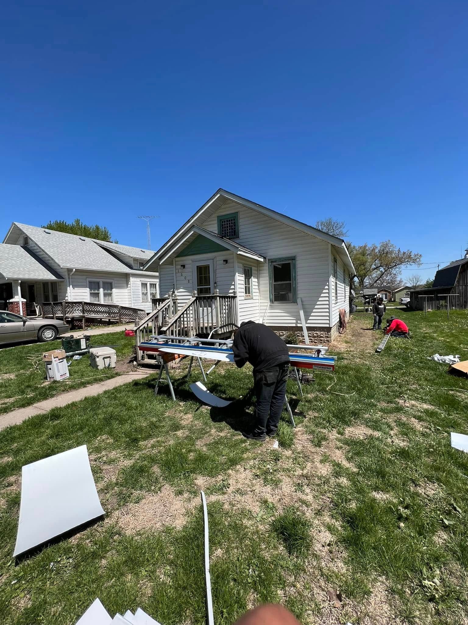 Person working in a messy front yard outside a small white house under a clear blue sky