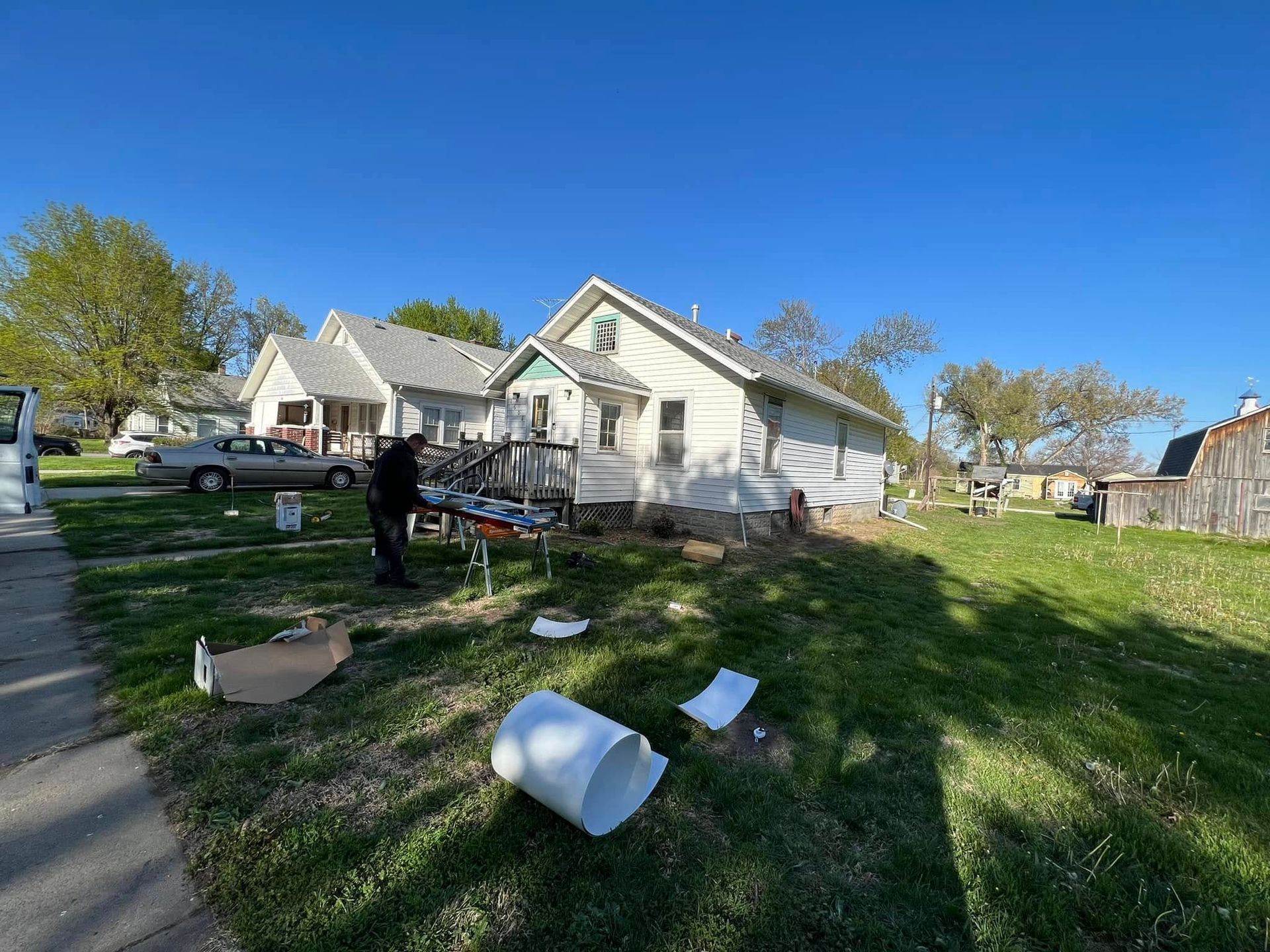 White house in a yard with scattered debris, a person near the porch, and trees under a blue sky.