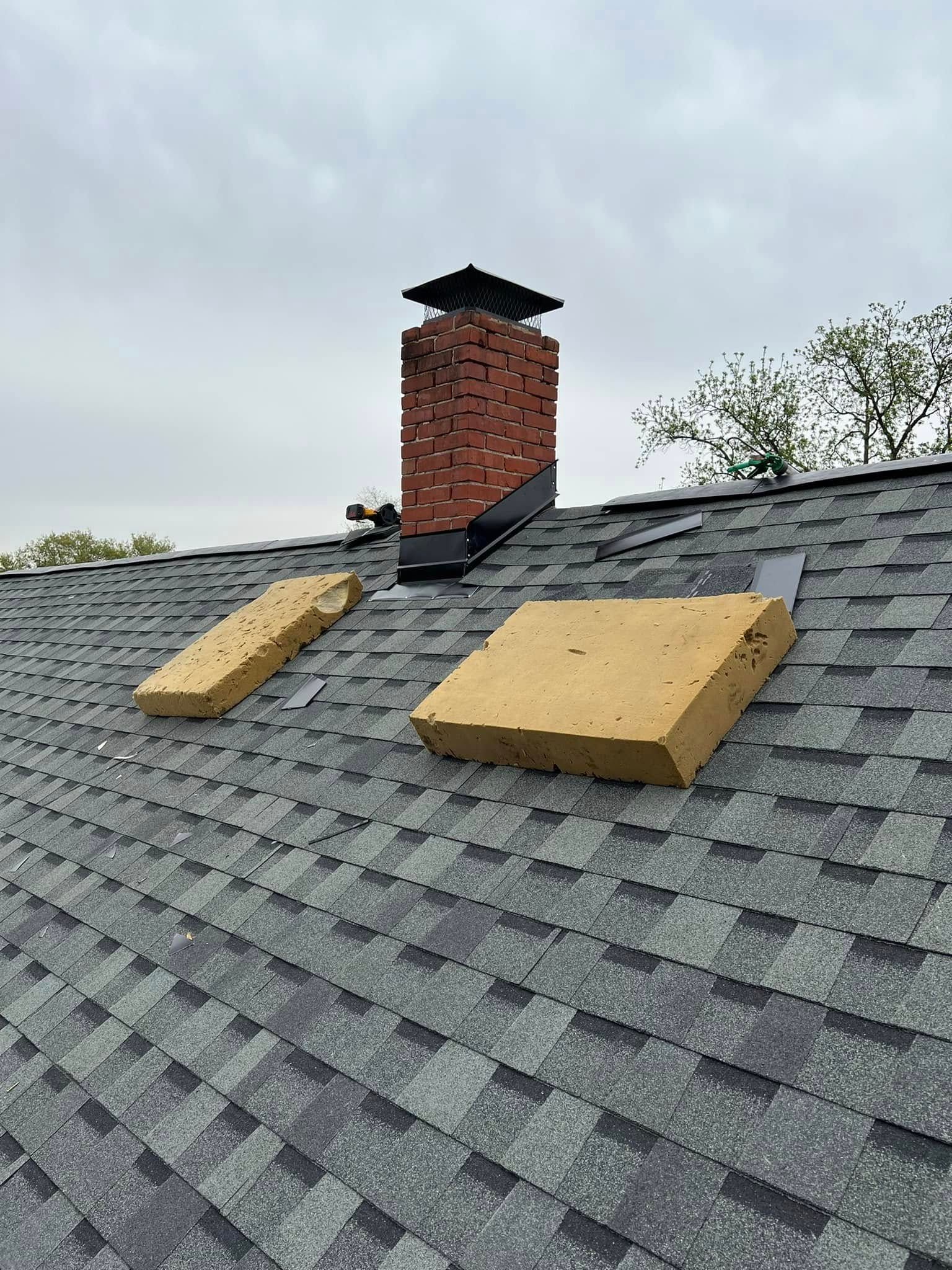 Rooftop with gray shingles, a brick chimney, and two tan foam pads near the chimney.