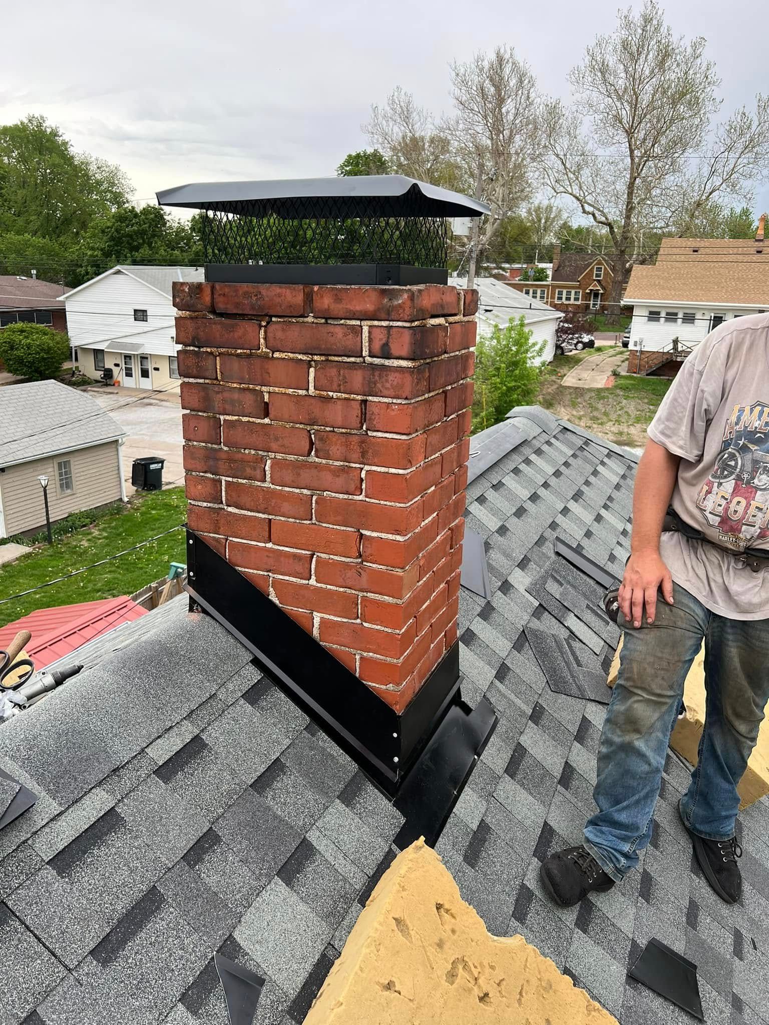Brick chimney on a gray shingled roof with a black metal flashing, next to a worker on the right.