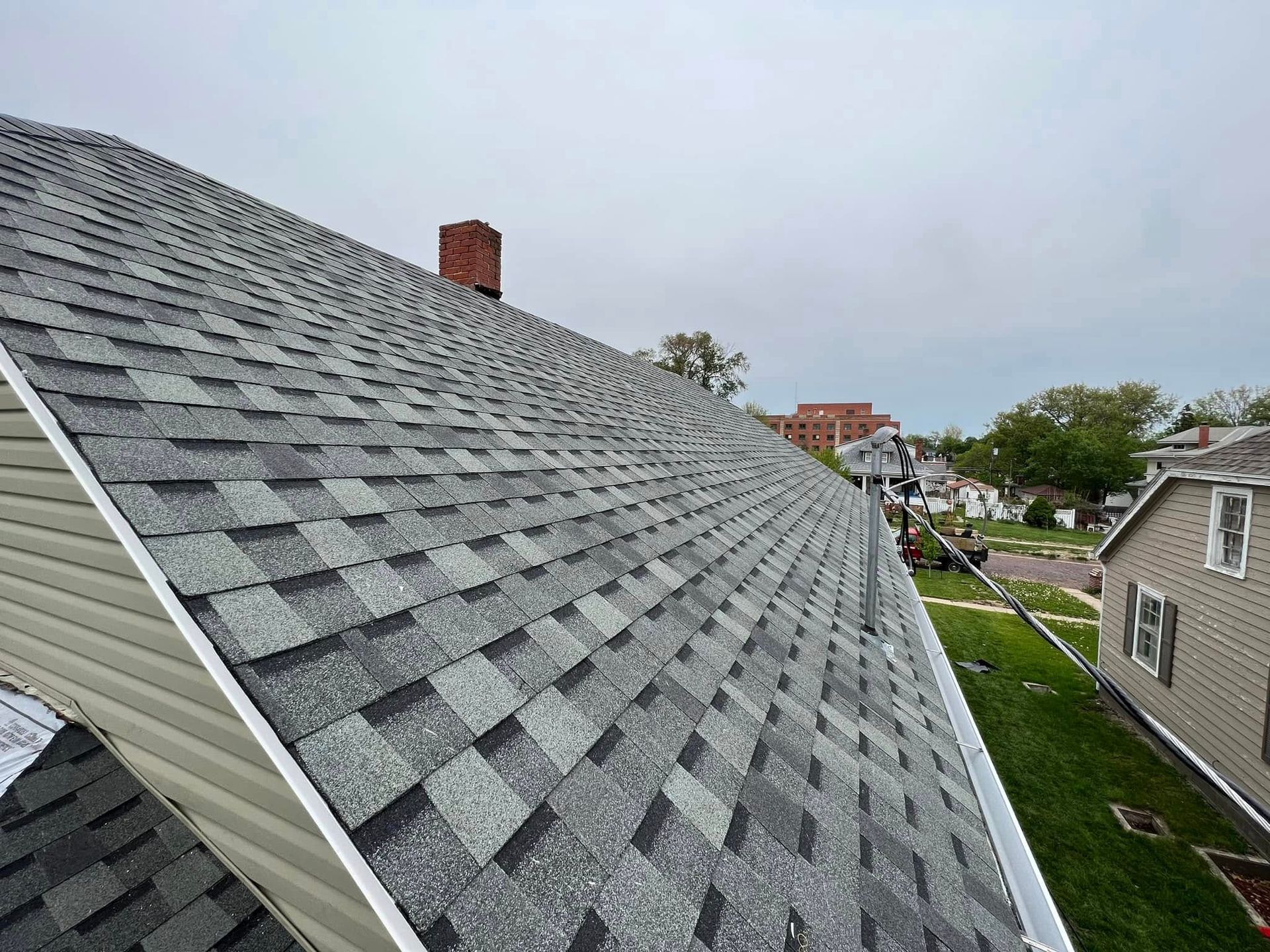Gray shingled roof on a house, viewed from above, with neighboring homes and cloudy sky in the background