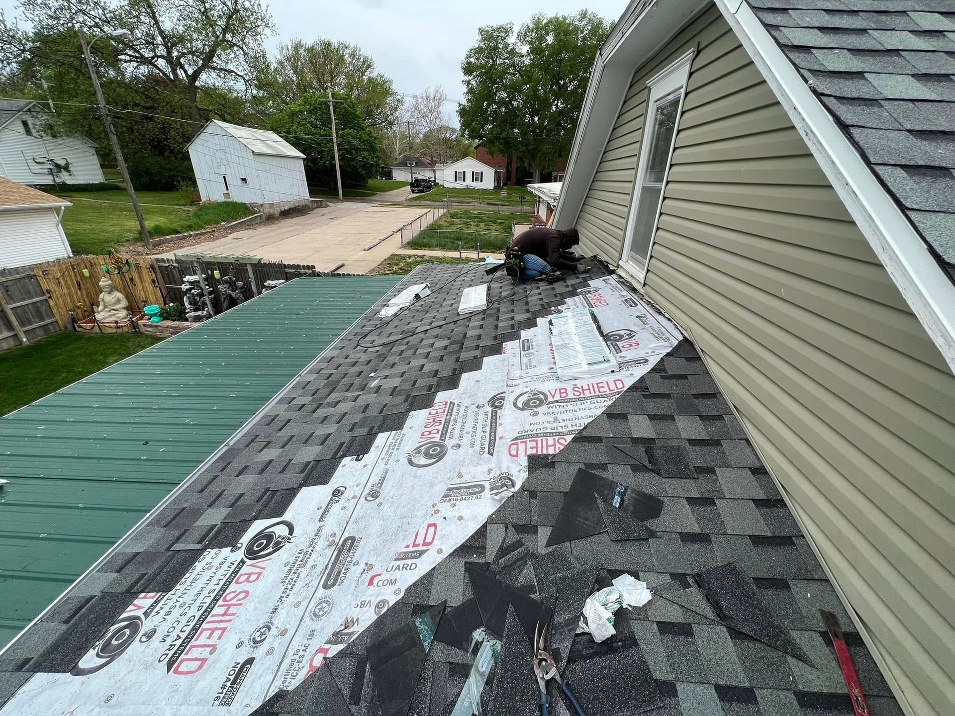 Roof under repair with shingles and underlayment beside a house, with a worker kneeling near the ridge.