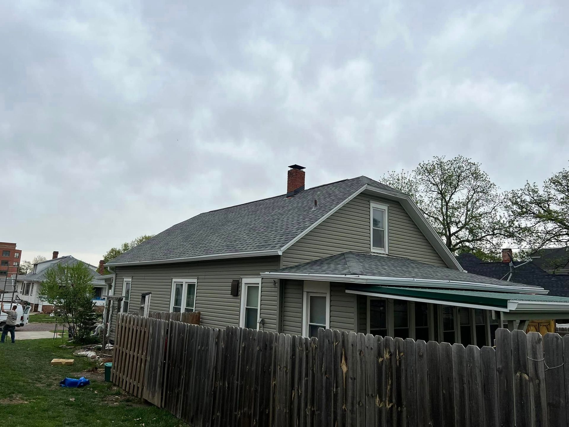 Weathered house with gray shingle roof, green awning, and wooden fence under cloudy sky