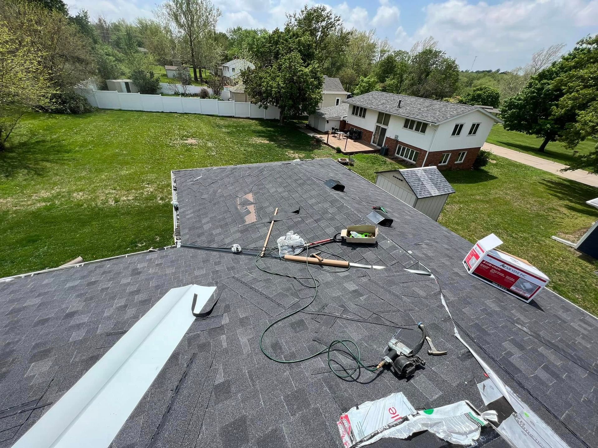 Roofer’s view of a shingled roof with tools, materials, and a house and yard below