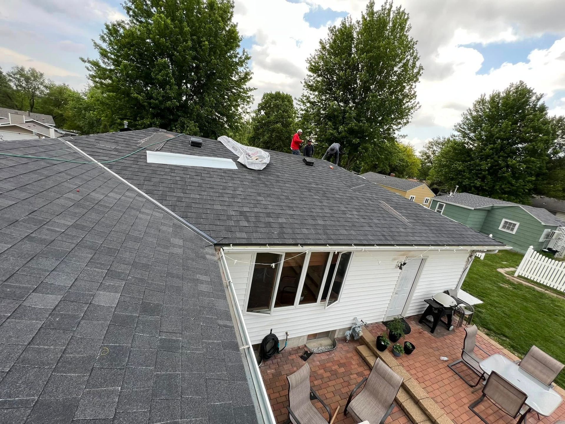Roof repair on a house with gray shingles, white siding, and a patio below, with two workers on top.