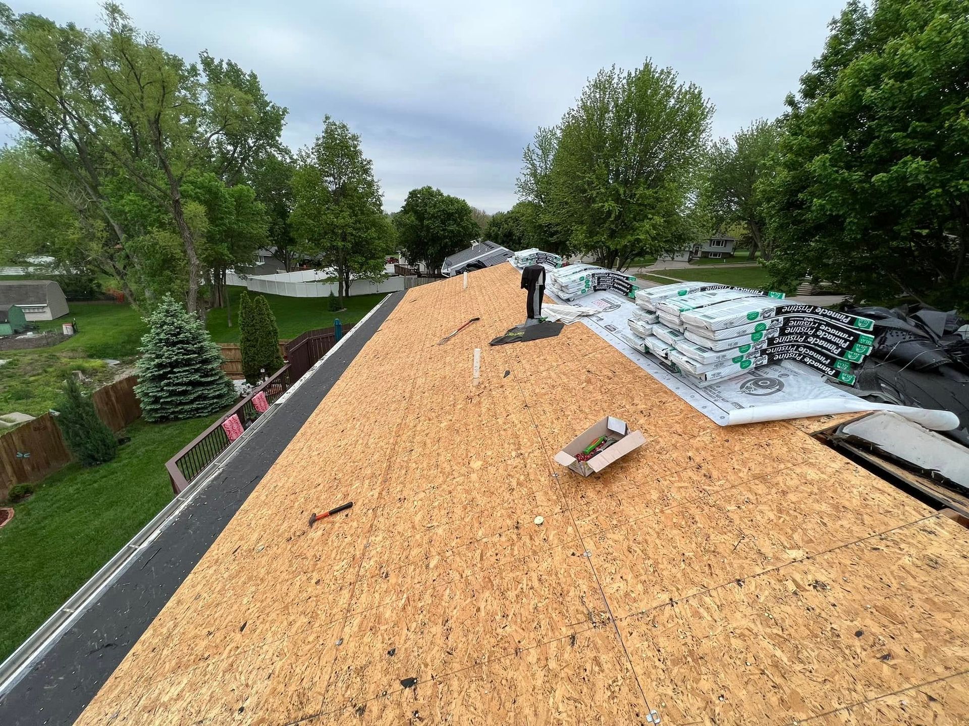 Roof under construction with plywood sheathing, workers, and stacks of materials beside trees