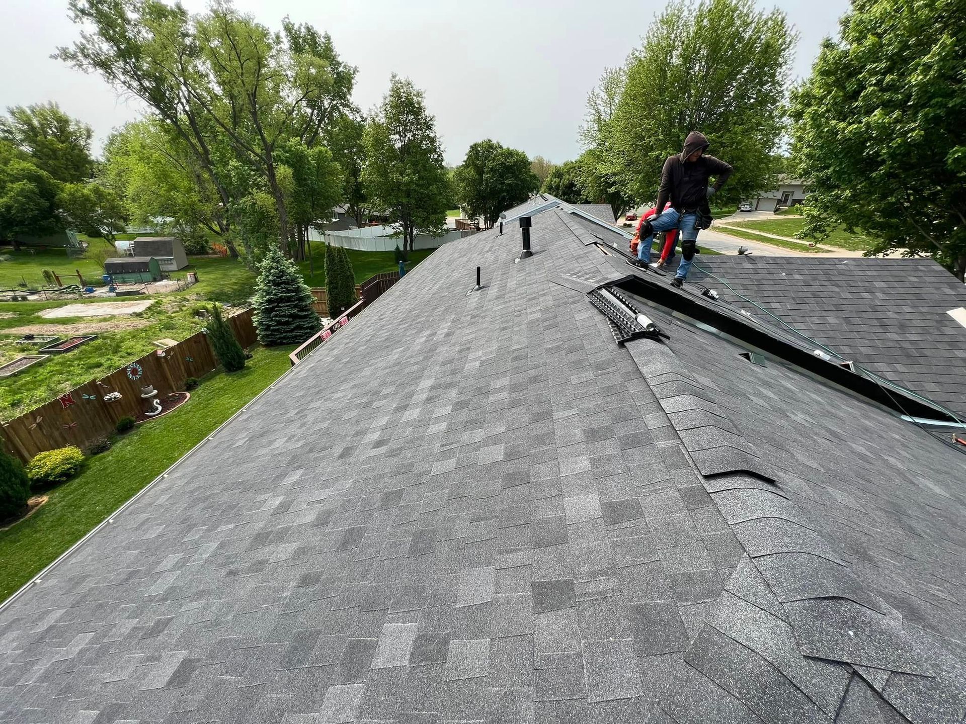 Workers repairing a gray shingle roof in a leafy residential area