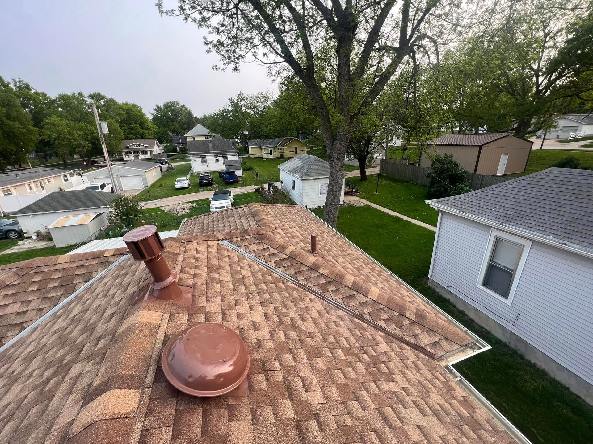 Rooftop view over suburban houses and trees, with tan shingles and a metal vent in the foreground