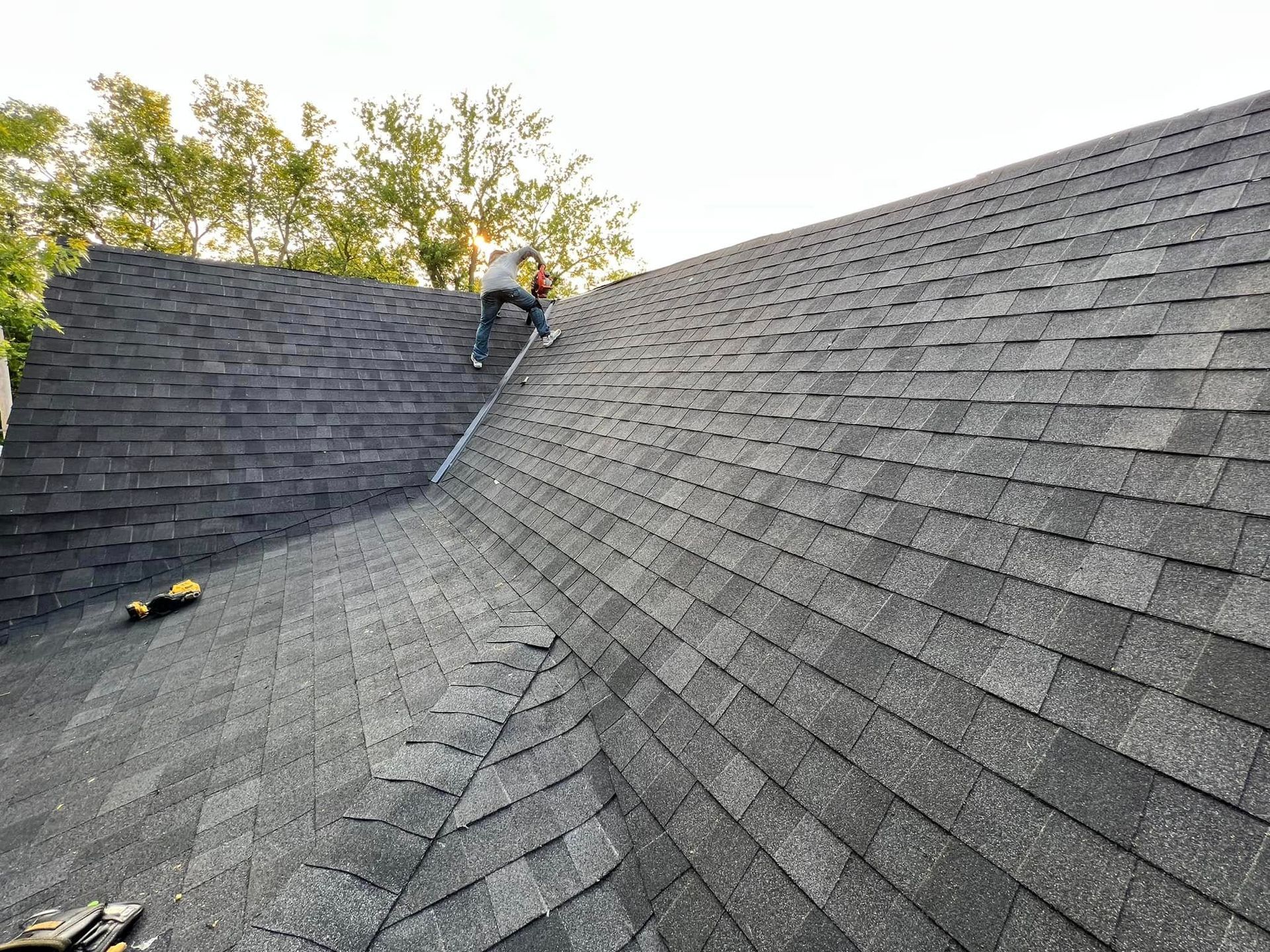 Worker on a steep gray shingle roof, with trees in the background.