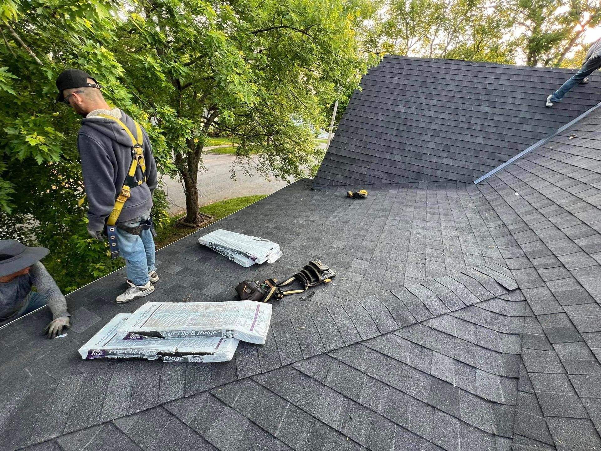 Two workers on a shingled roof with roofing materials and tools near trees.