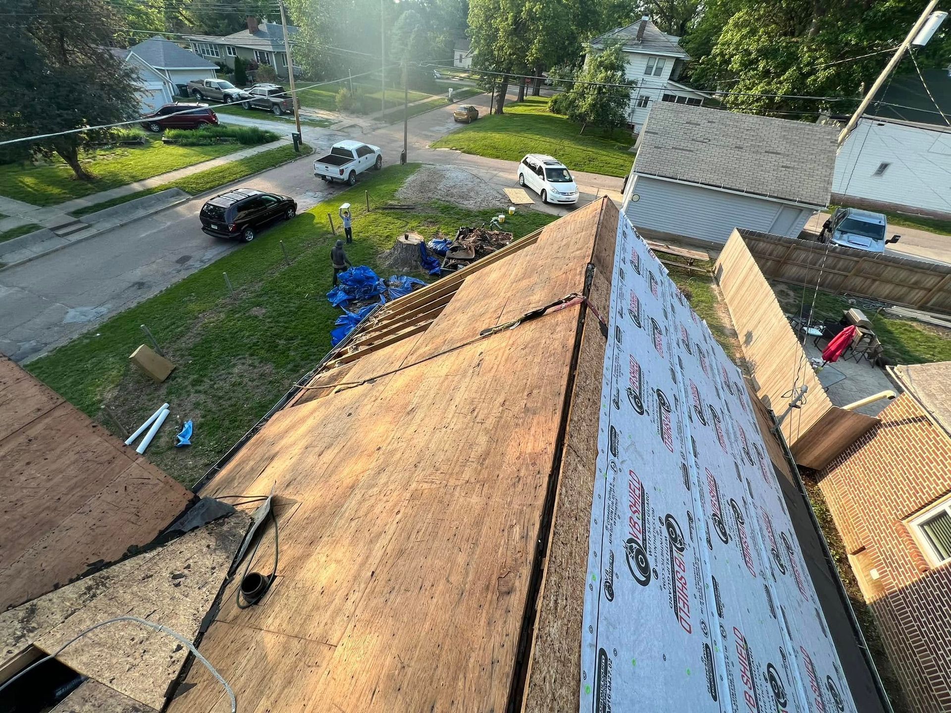 Rooftop view of a neighborhood street with parked cars, trees, and roof shingles under repair