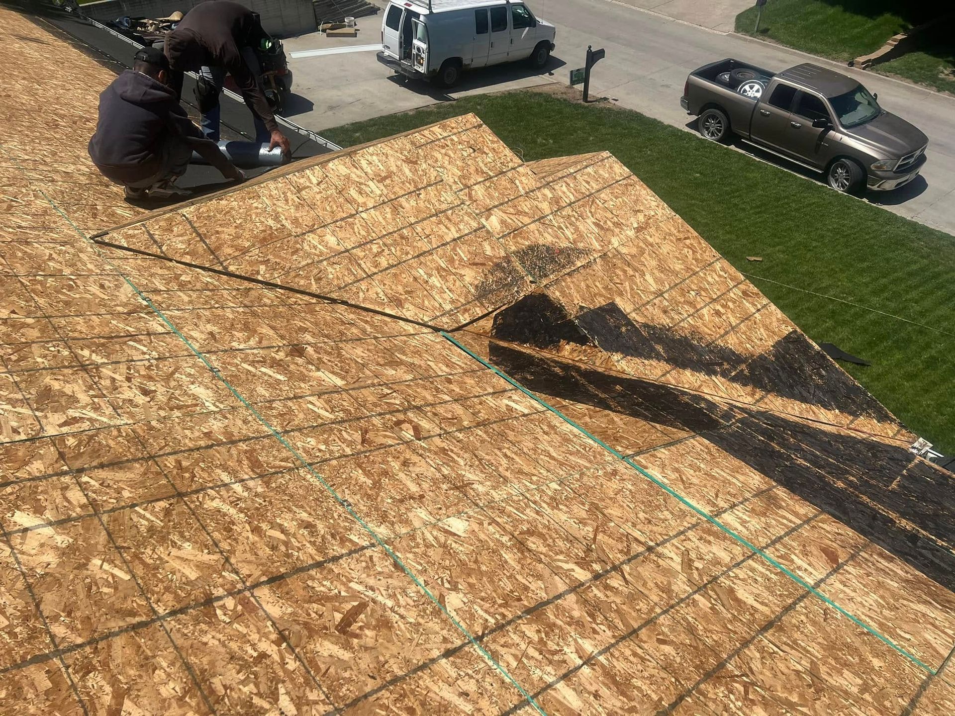 Workers installing roofing plywood on a house roof, with tools and trucks below.