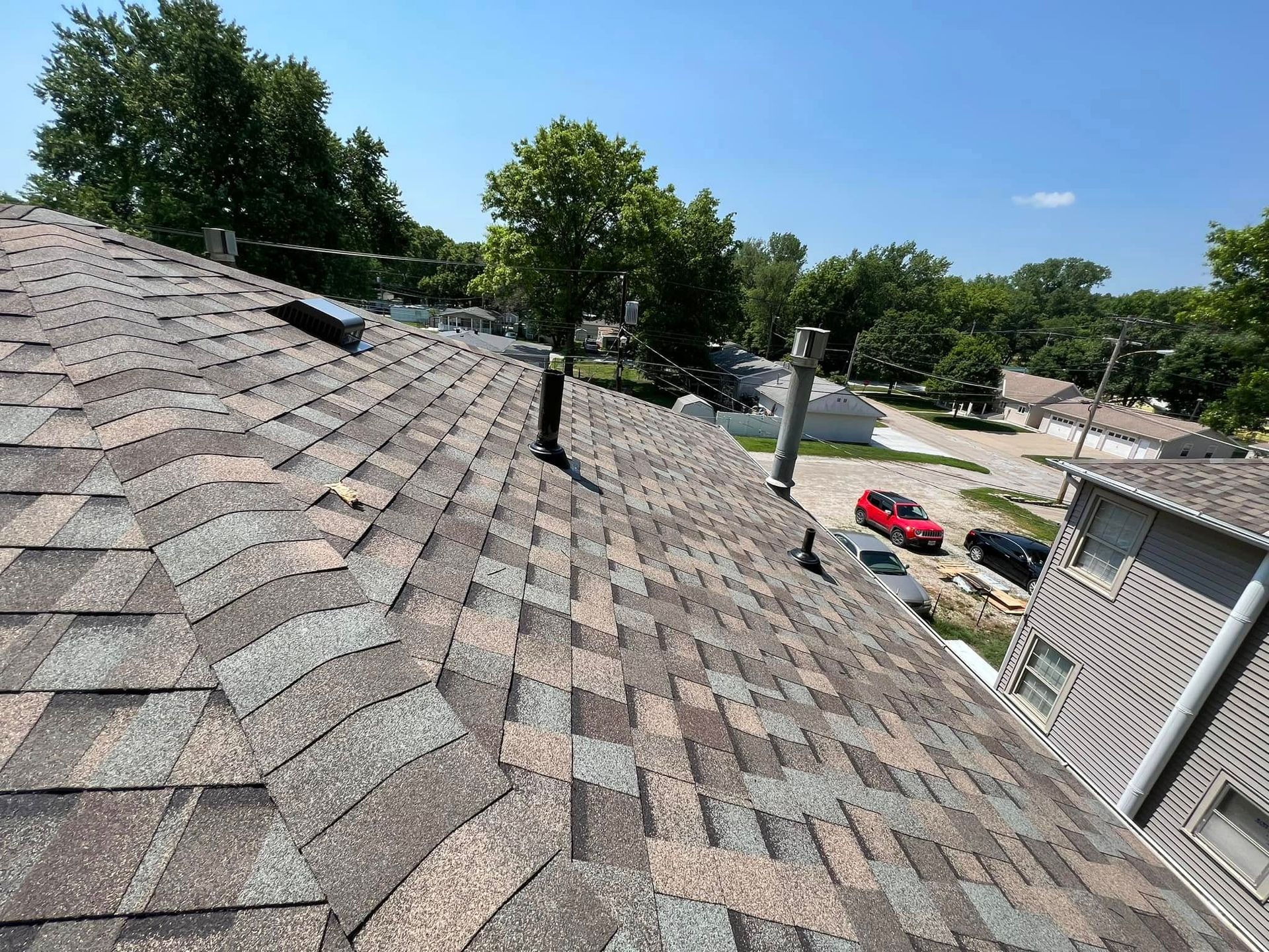 Roof shingles with a chimney and suburban houses, trees, and parked cars under a blue sky