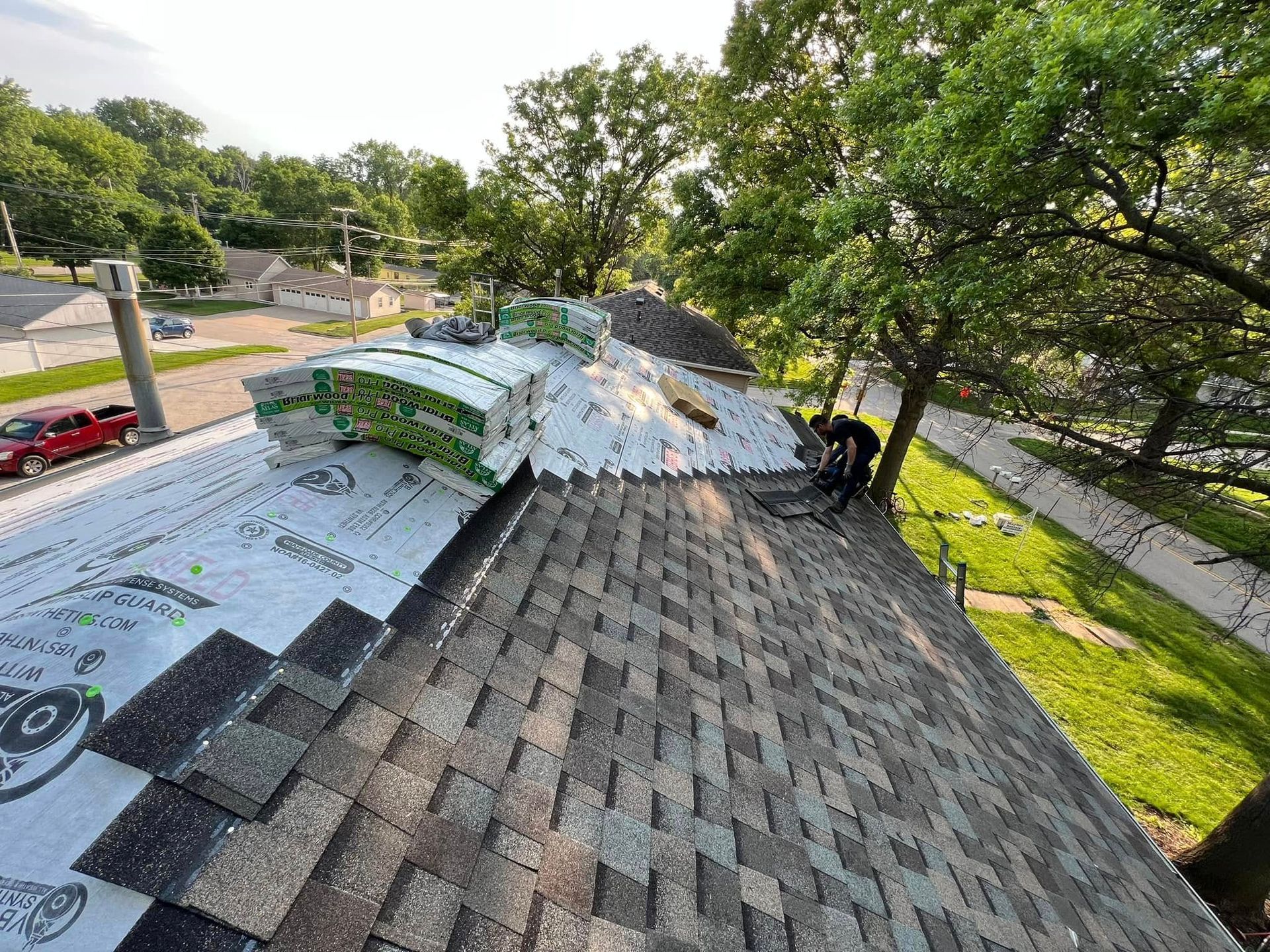Workers installing shingles on a residential roof beside trees and a road