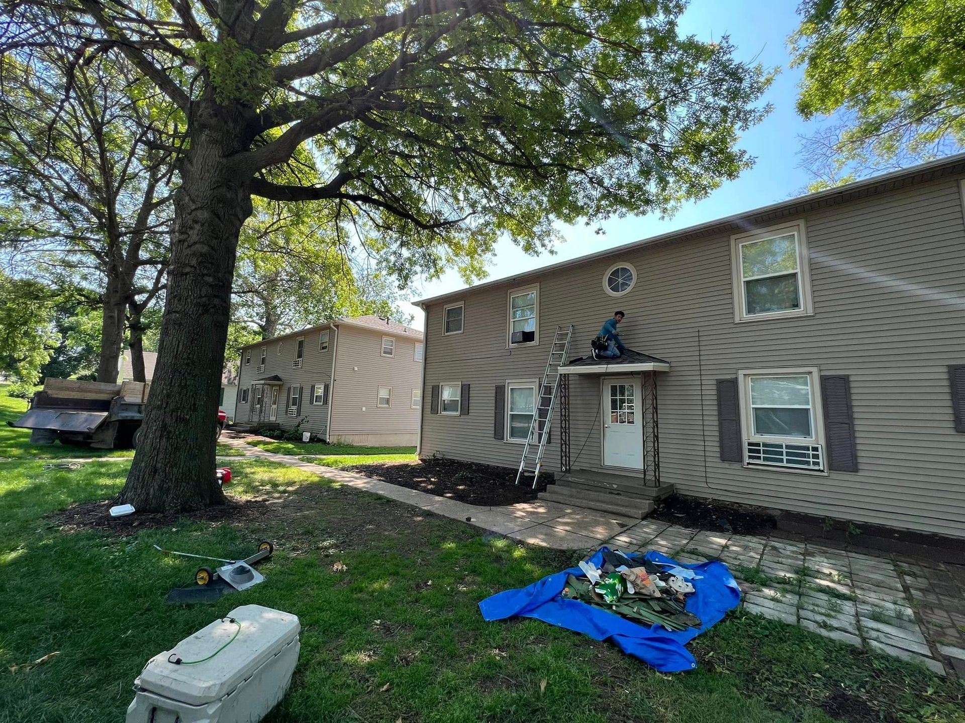 Residential building with large tree, debris tarp, and lawn equipment in front yard