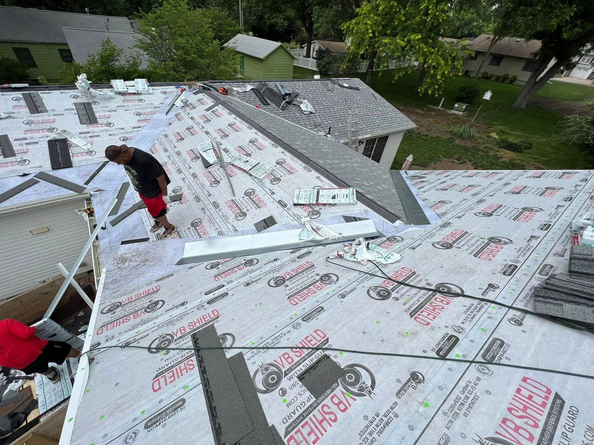Workers installing white roofing underlayment on a sloped roof beside trees and houses