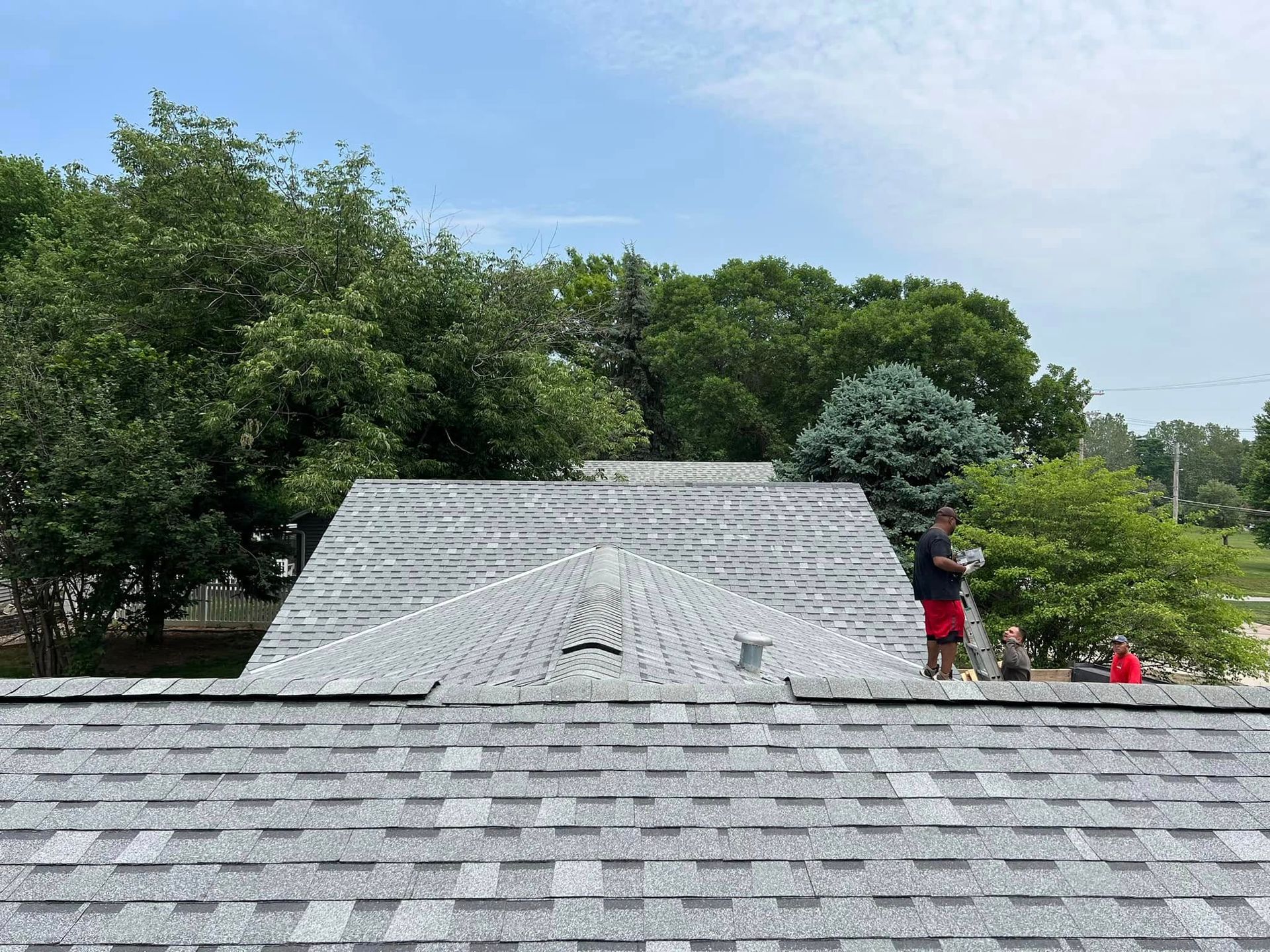 Shingled rooftop in a leafy park, with a few people walking in the background under a blue sky.
