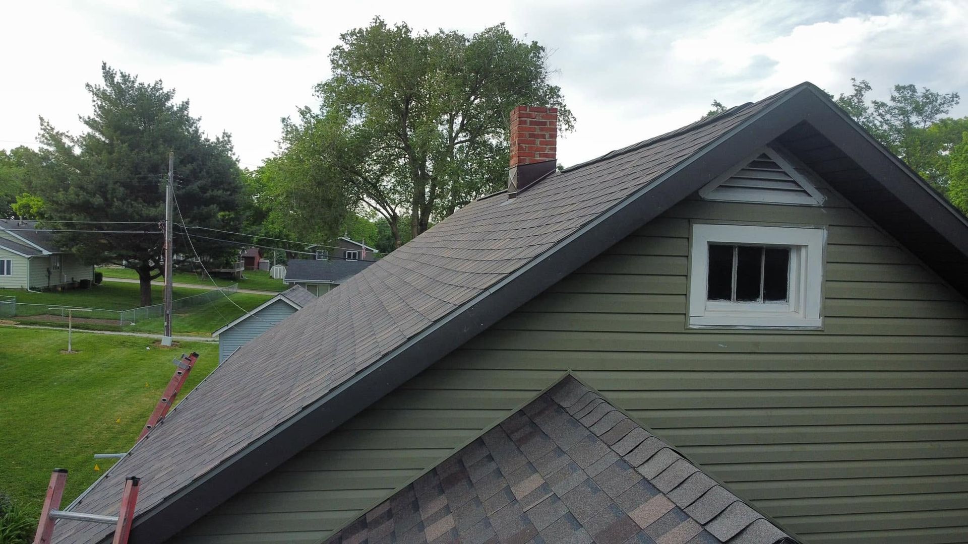 House roof with gray shingles, dormer window, and brick chimney beside trees and a grassy yard