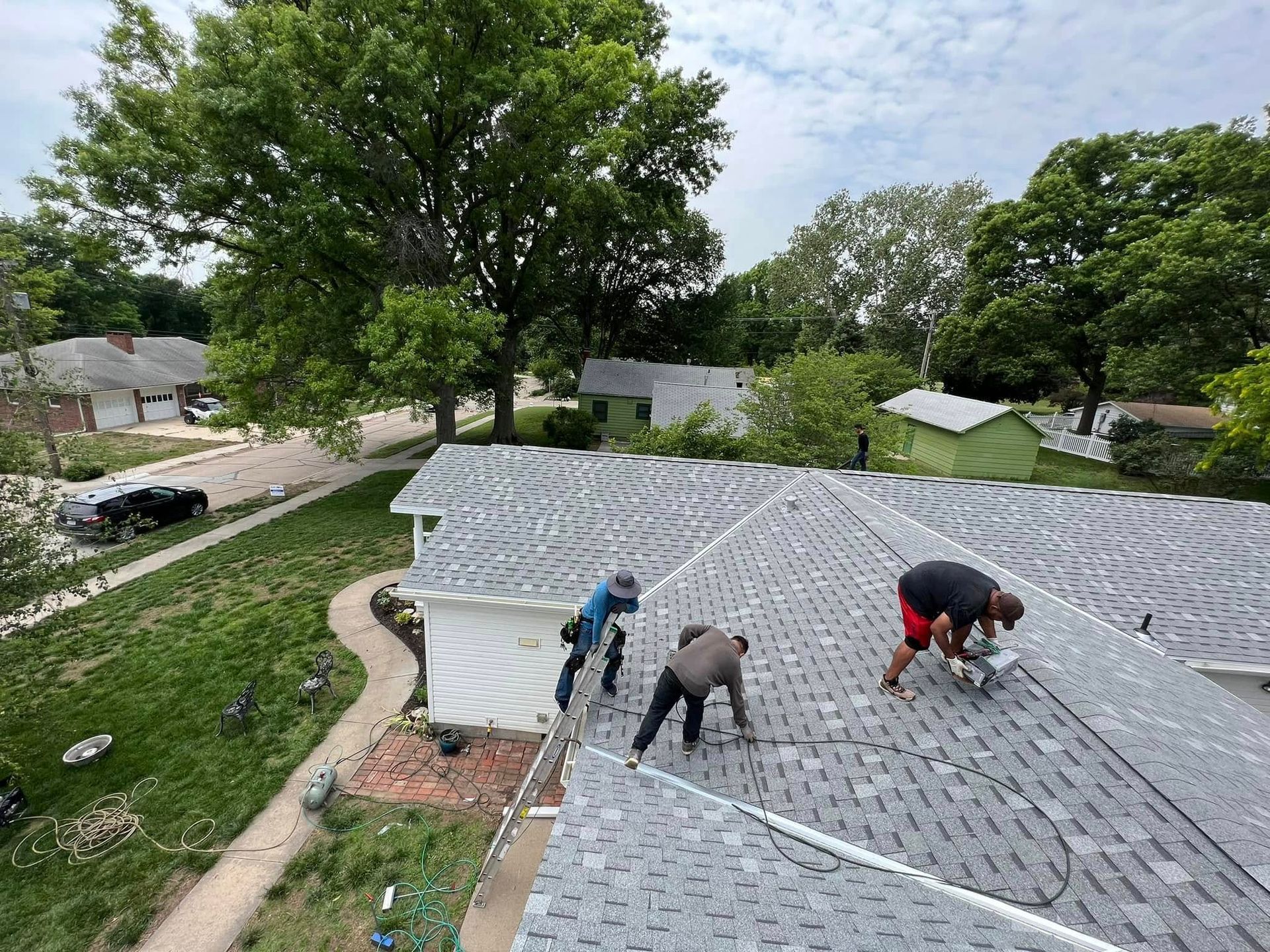 Workers installing gray shingles on a curved roof beside trees and a grassy yard