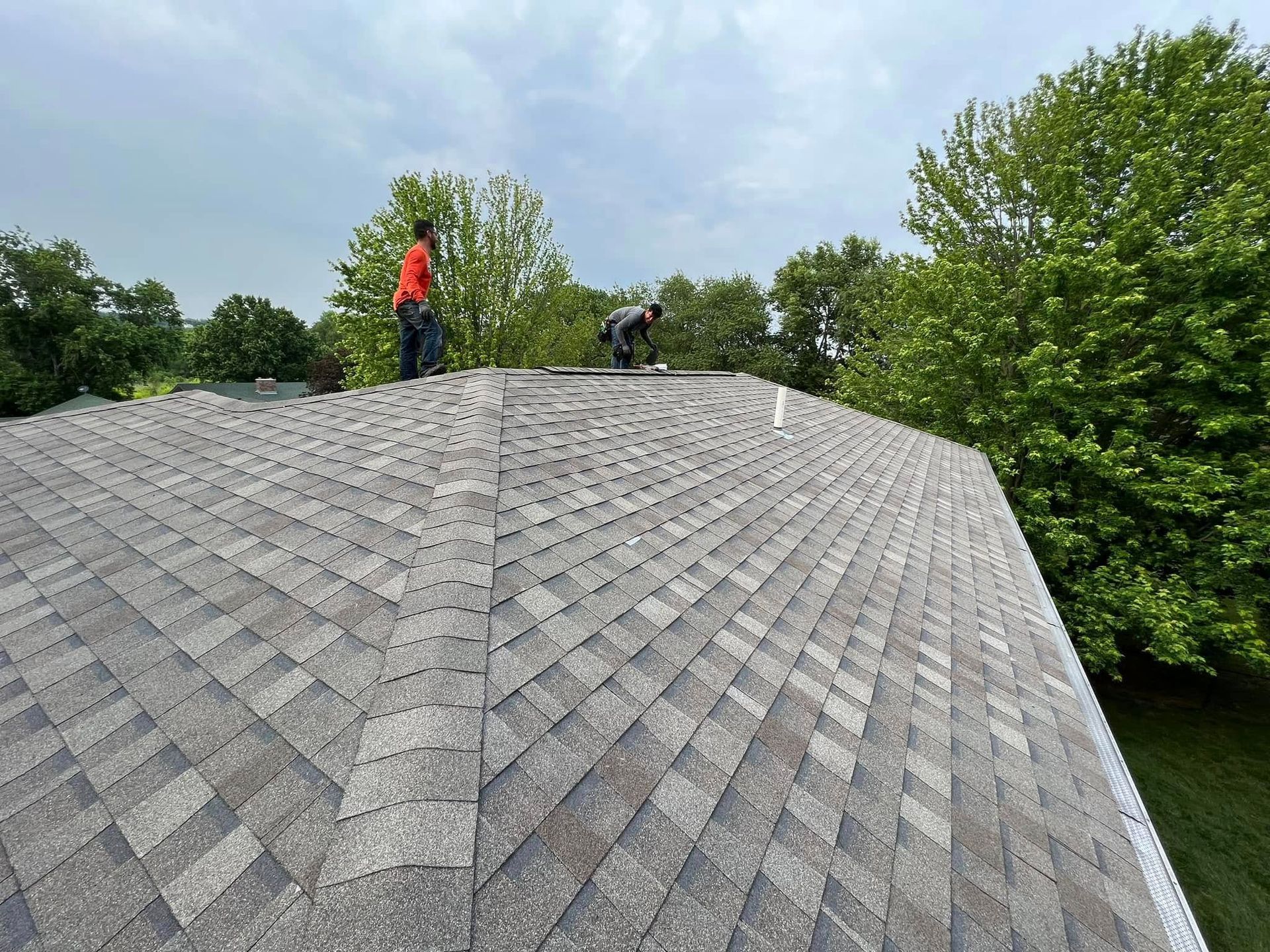Two workers inspecting a gray shingled roof under a cloudy sky, surrounded by trees