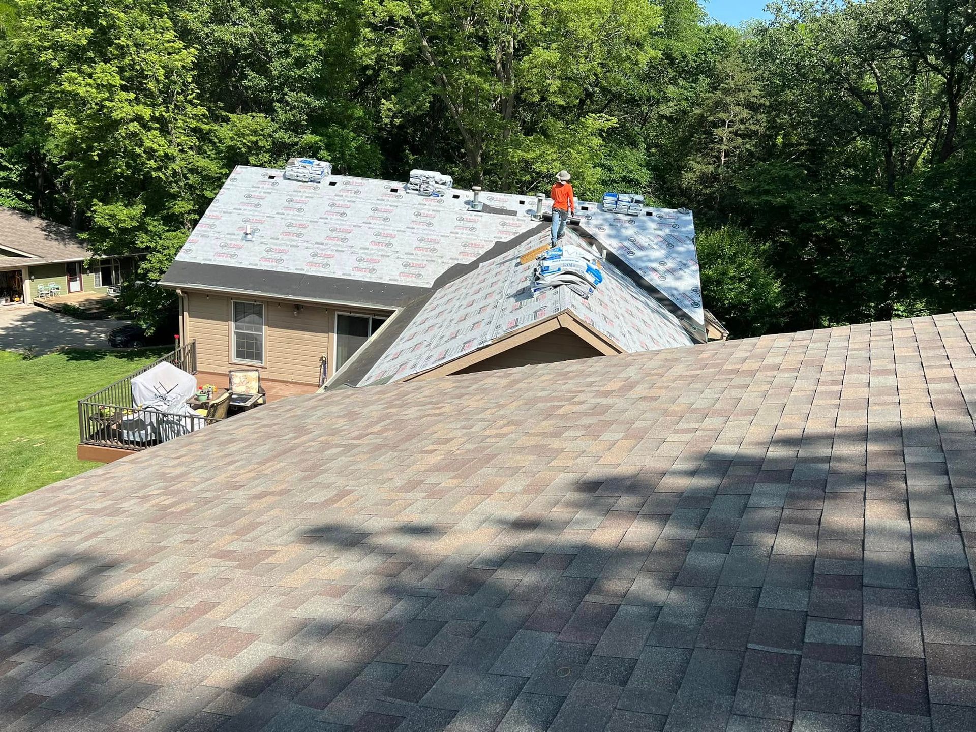 Roofers working on a gray shingled roof beside a wooded house during repairs