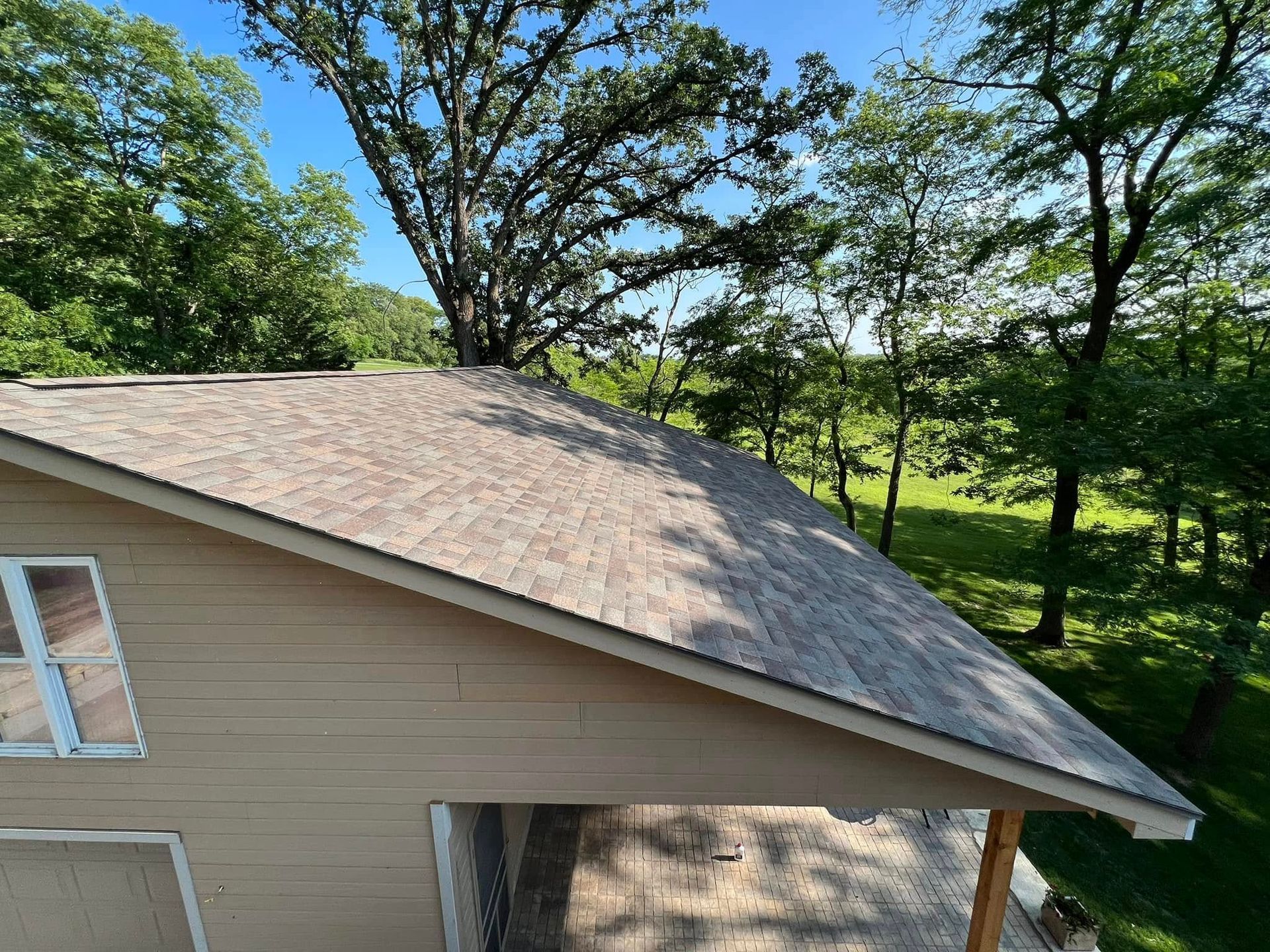 Tan house with gray shingled roof beside leafy green trees on a sunny day