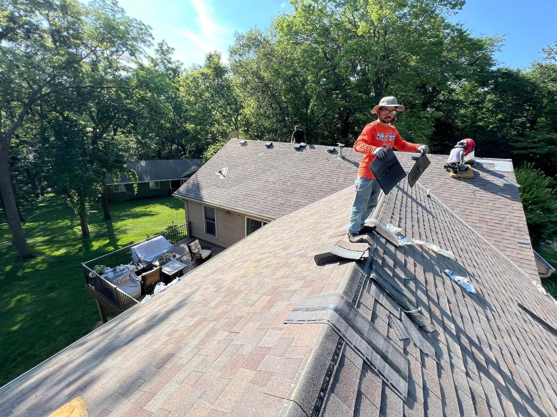 Workers roofing a house, one standing on a sloped gray roof near trees.