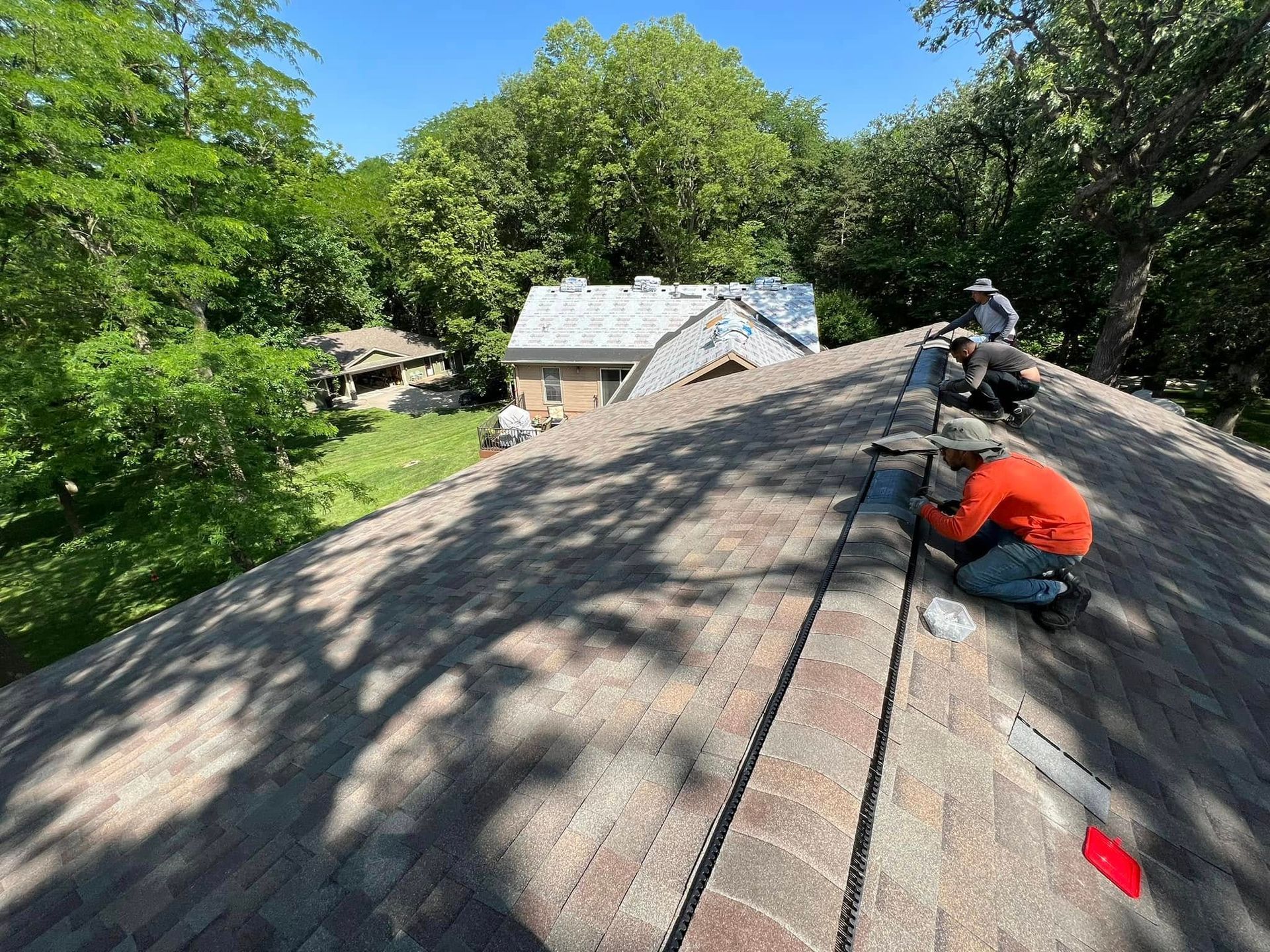Two roofers on a shingled roof beside trees, working with tools on a sunny day.
