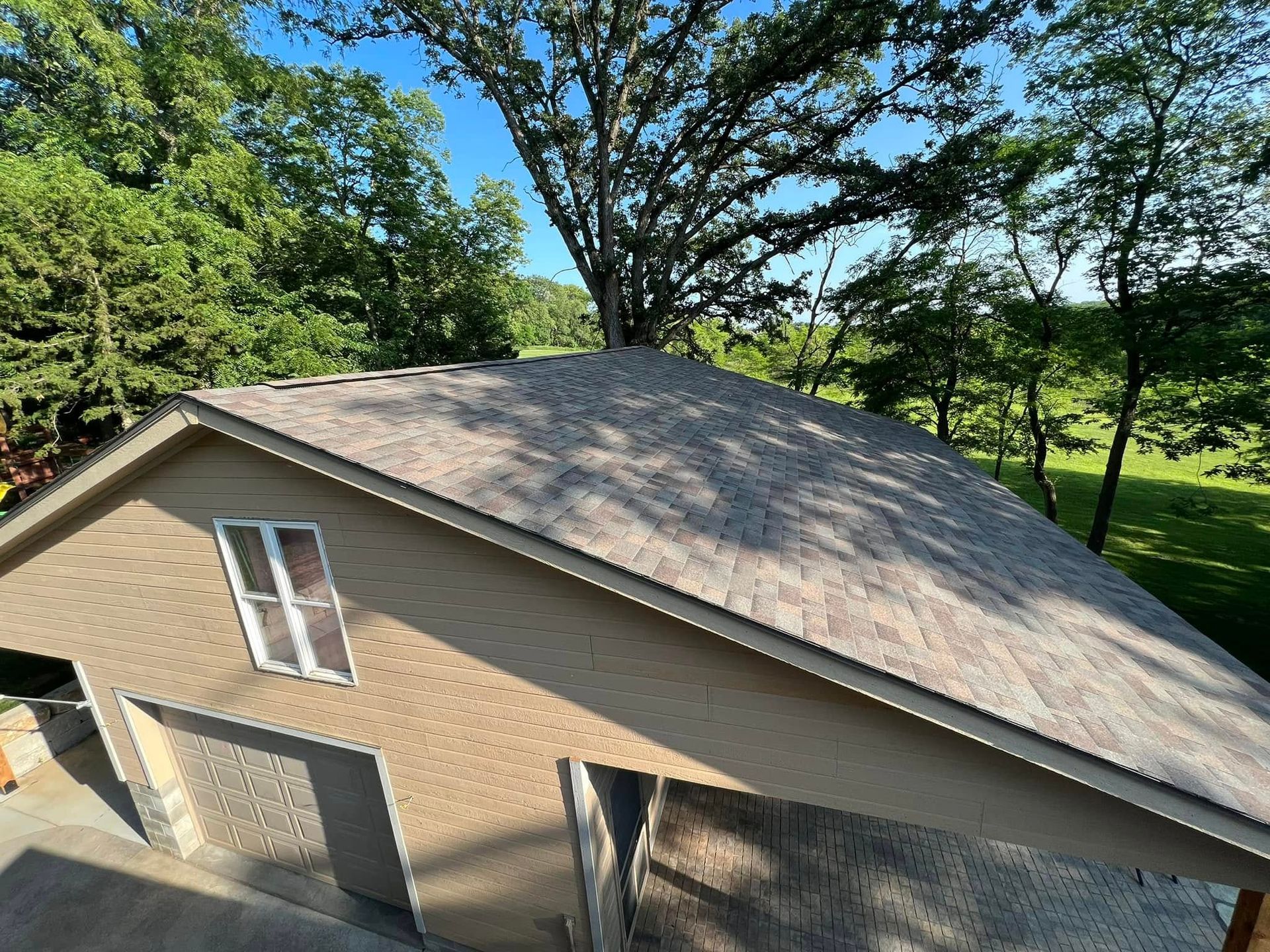 Brick house with gray shingle roof beside green trees on a sunny day