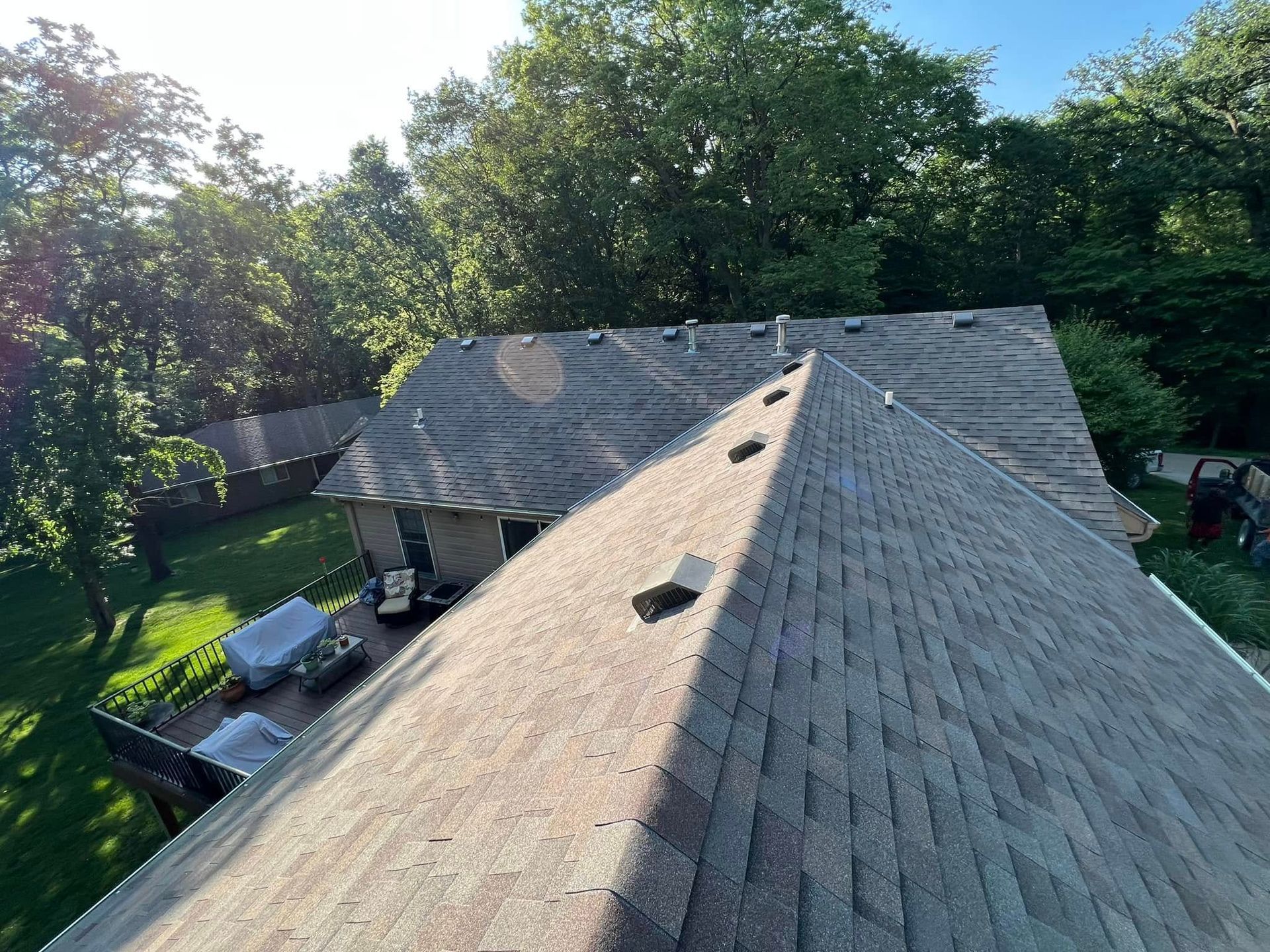 Rooftops with gray shingles and skylights beside a green backyard and trees in sunlight