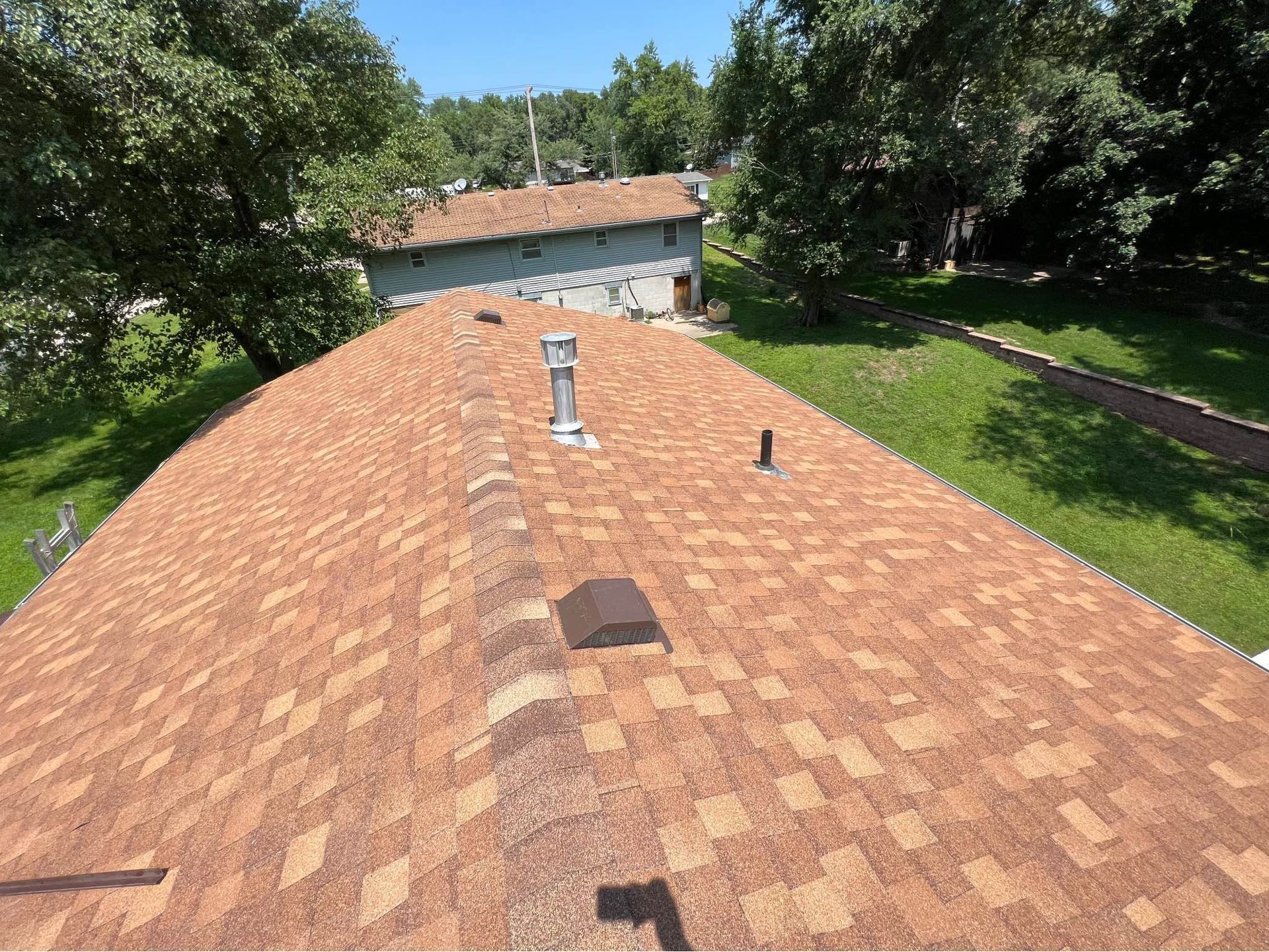 View from a shingled roof with vents, surrounded by trees and a smaller building in the background