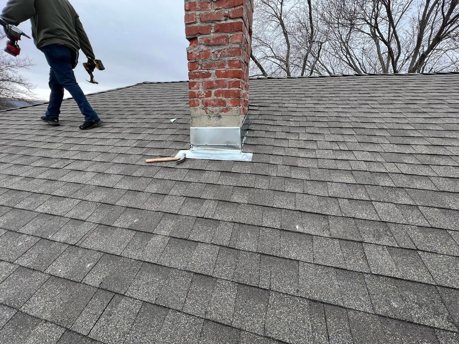 Worker walking on gray shingled roof beside brick chimney with flashing