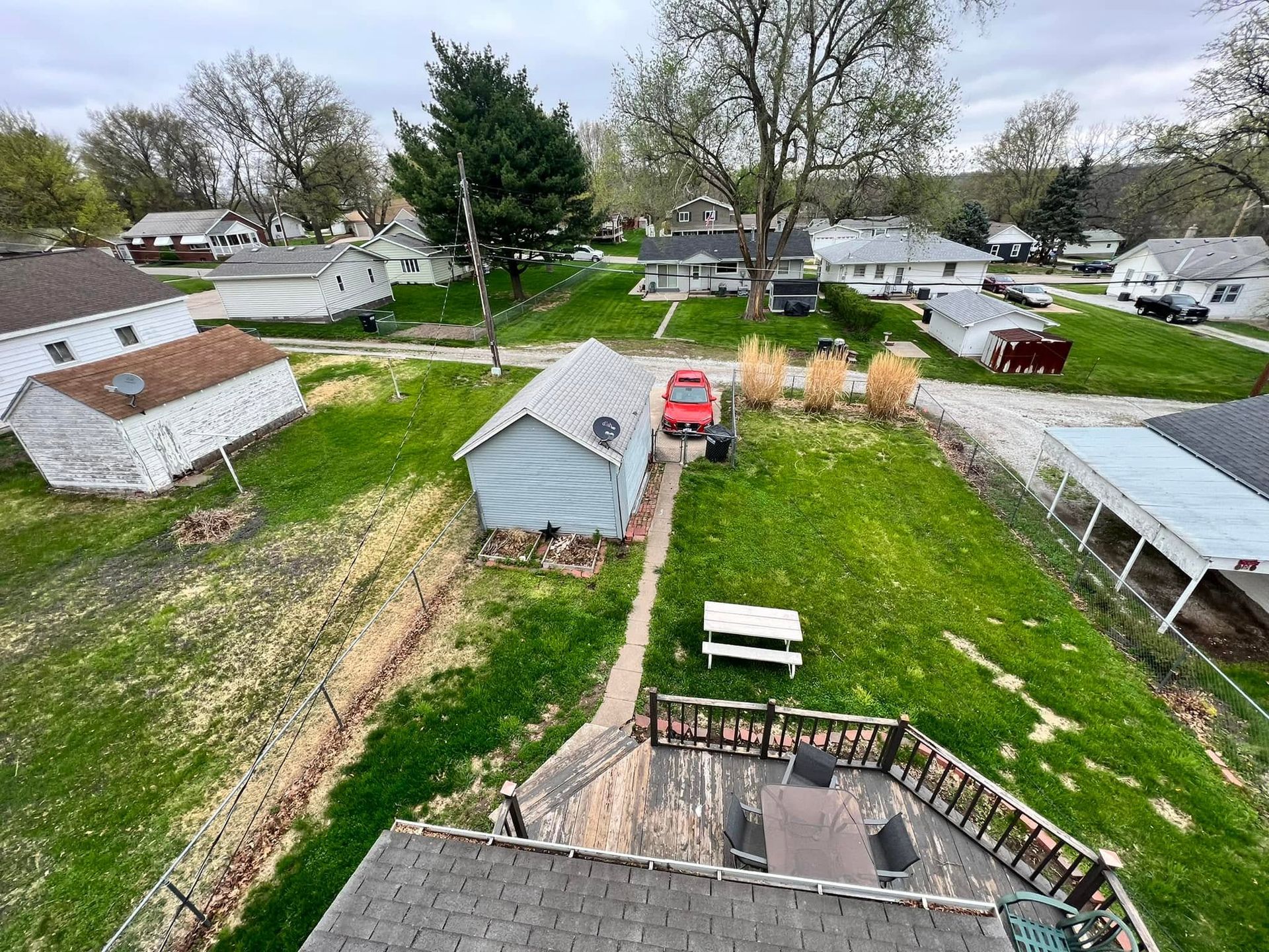 Overhead view of a grassy residential yard with sheds, a red car, and neighboring houses.