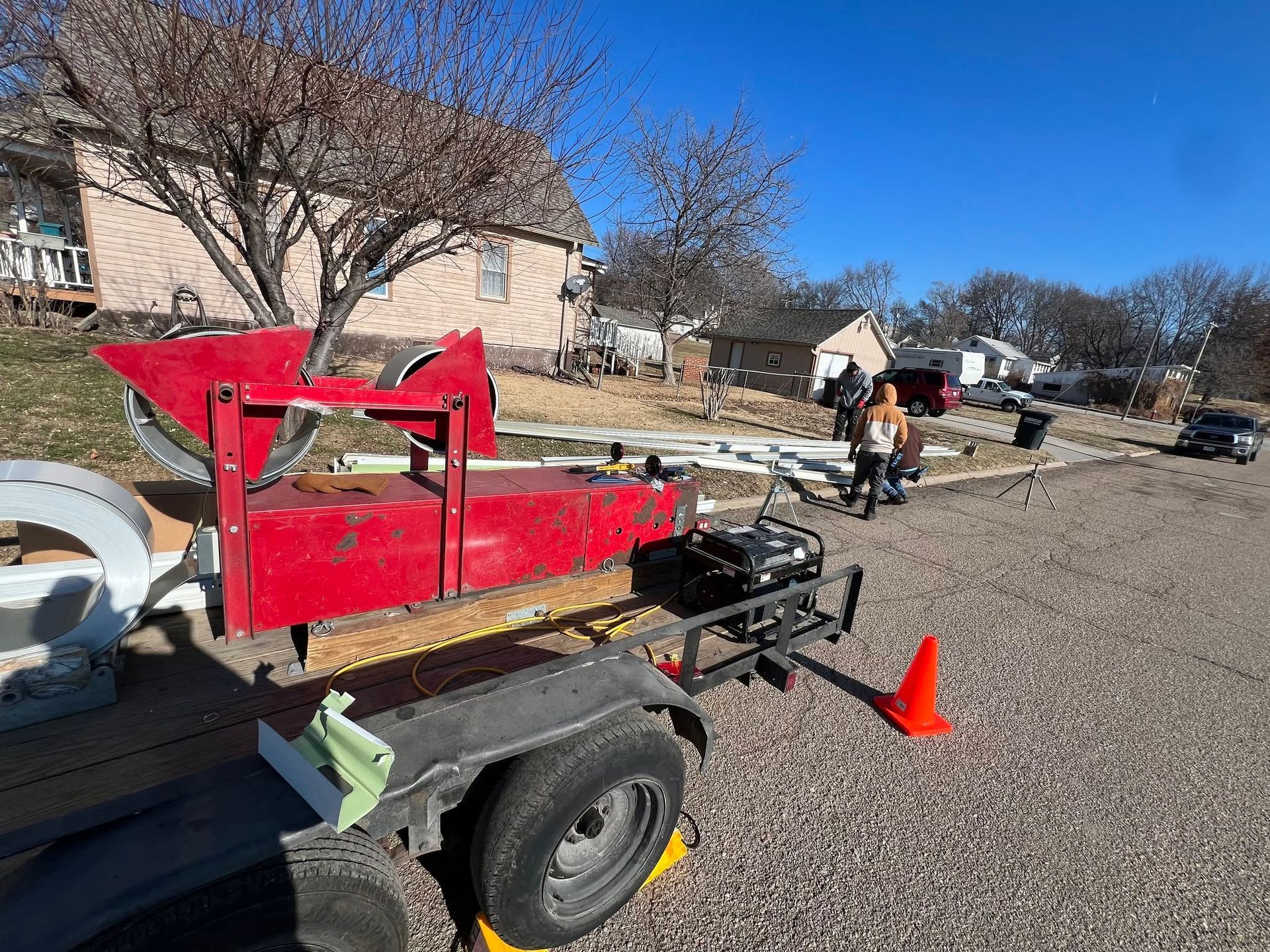 Red farm trailer and equipment parked on gravel with an orange cone in a yard under a blue sky.