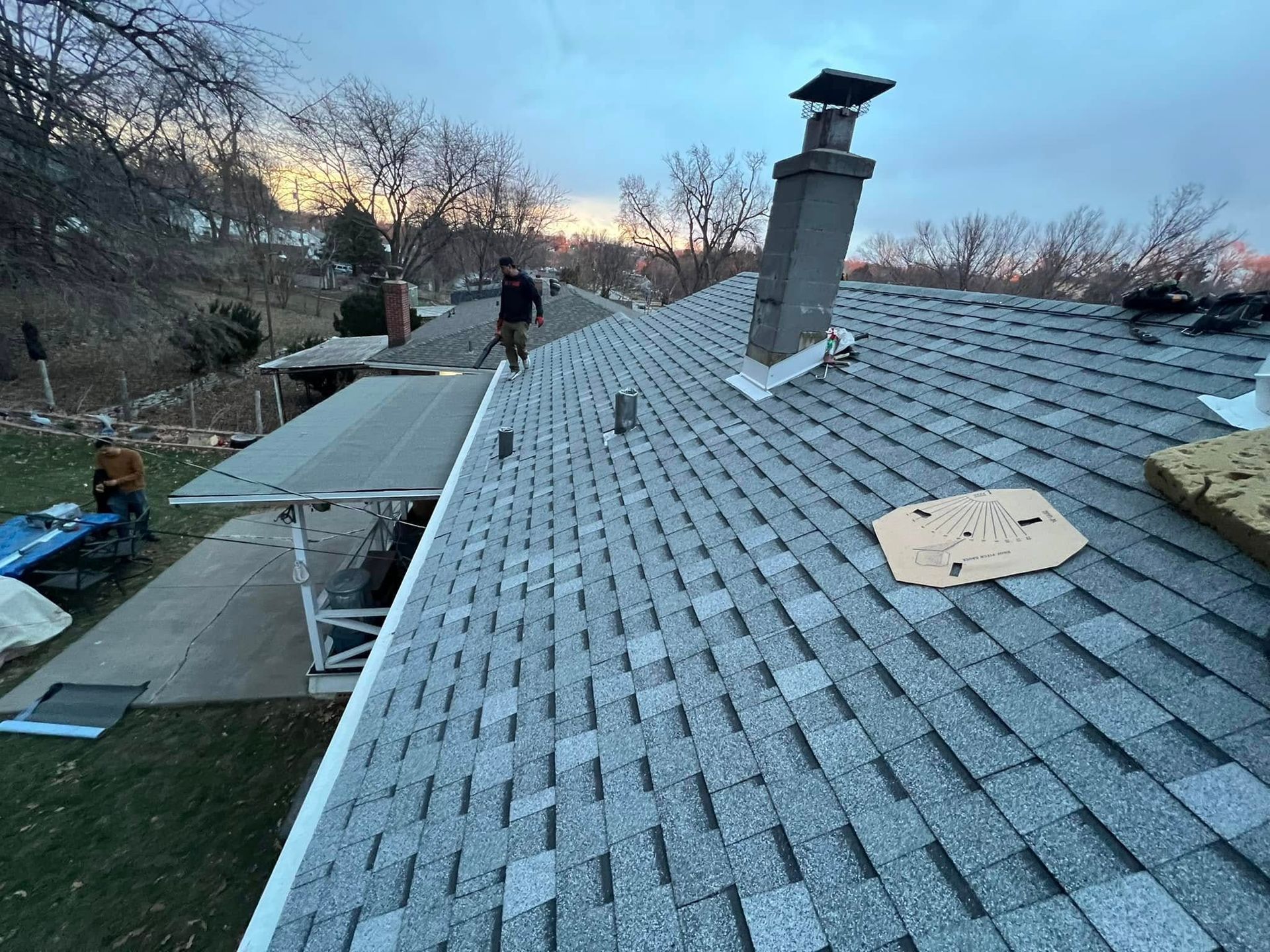 Rooftop with gray shingles, chimney, and a worker near the left edge at dusk